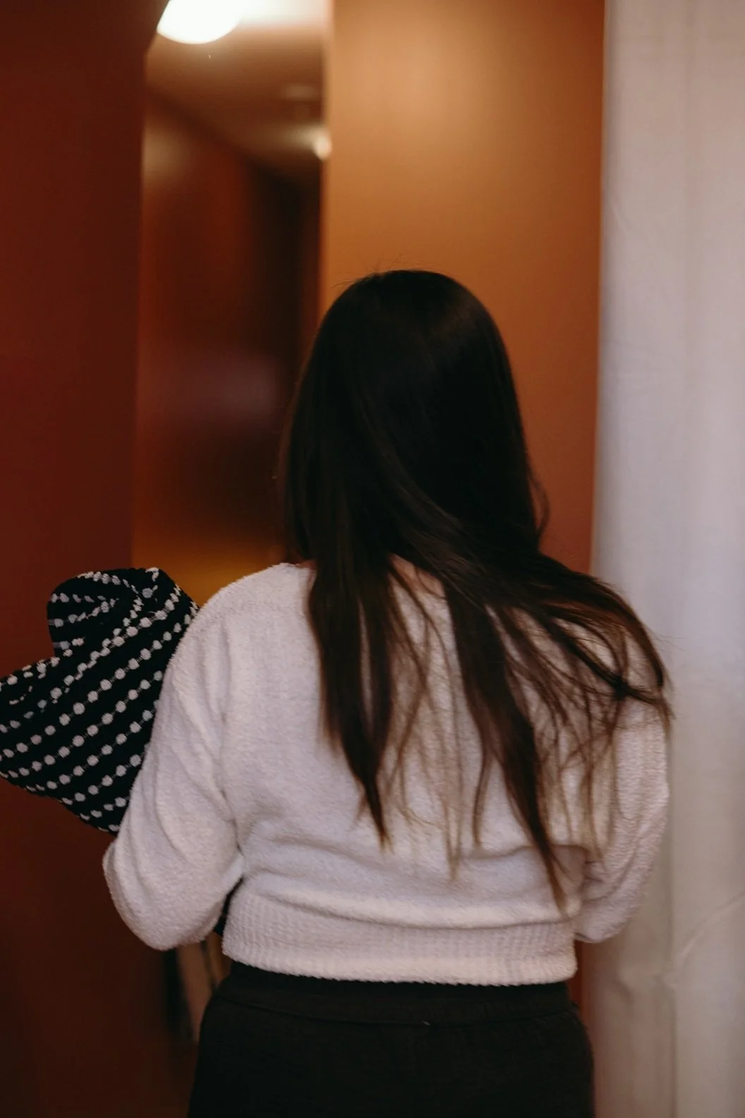 woman with long dark hair wearing a white sweater and black pants, walking down a corridor with wooden walls and a white curtain on the right side.