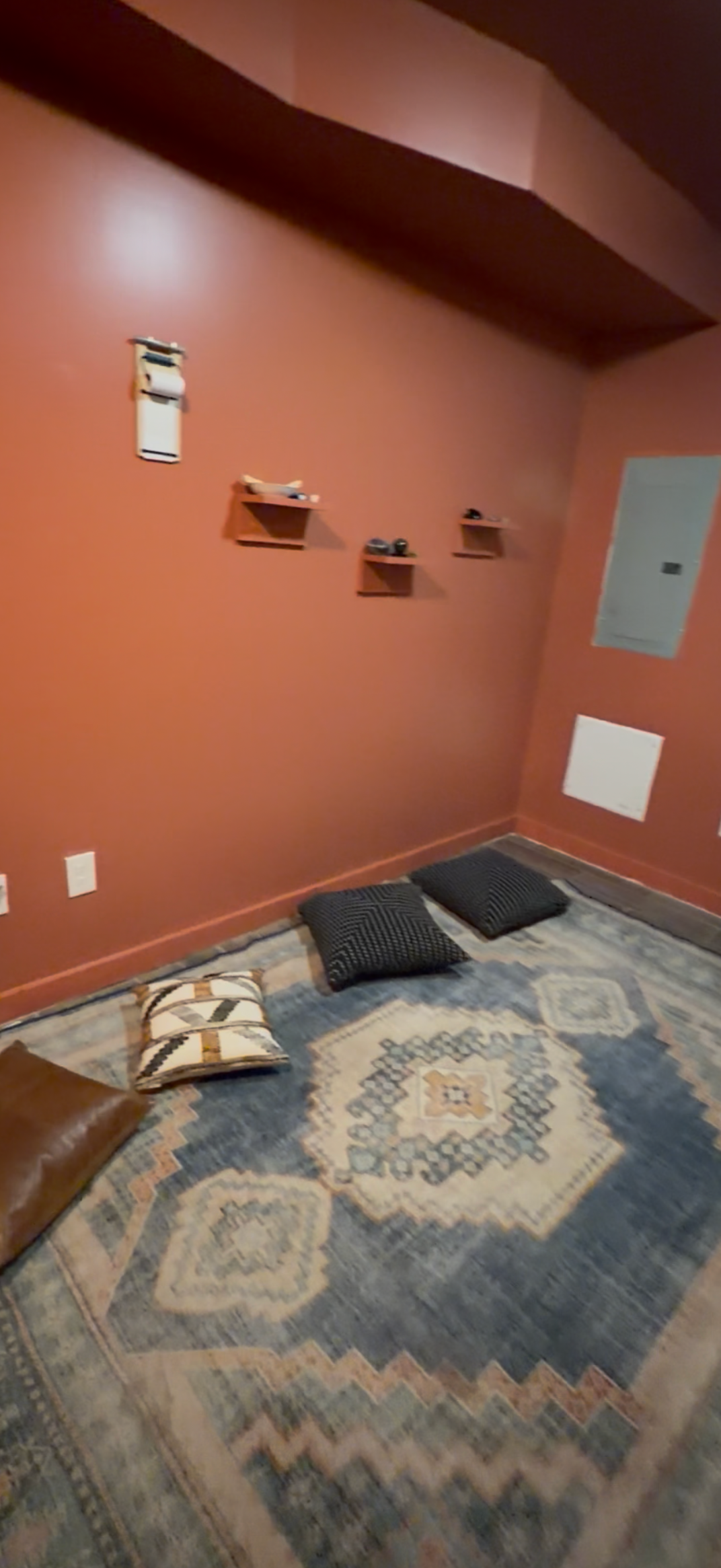 Photo of a cozy corner with a patterned rug, three throw pillows arranged on the floor, a pink painted wall with small wall-mounted shelves holding small objects, and an electrical panel.