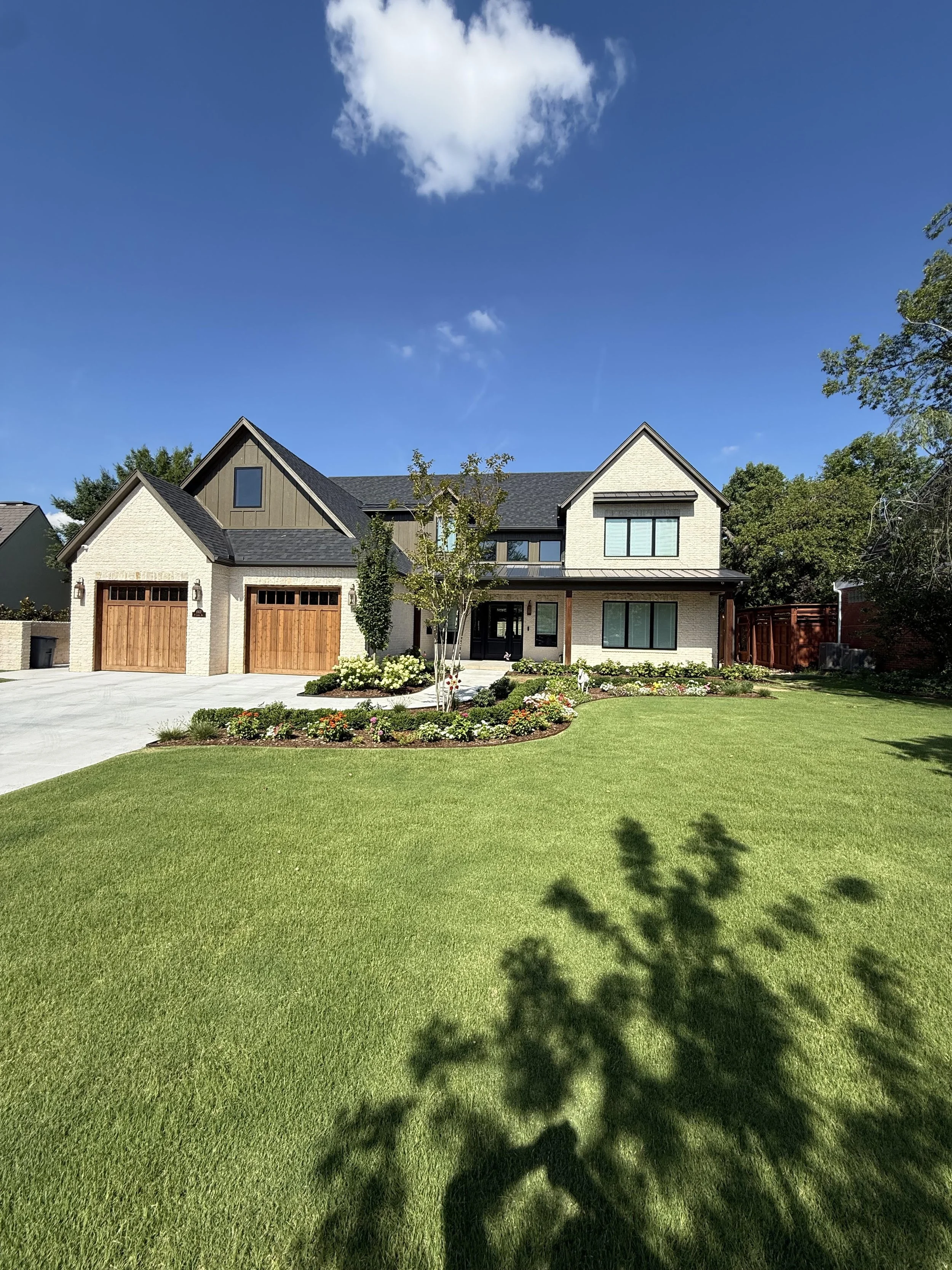A modern two-story house with a large front yard, trimmed green lawn, and a flower bed near the entrance. The house has a combination of white brick and darker siding, with two wooden garage doors and a driveway. Trees and a blue sky with a few clouds are in the background.