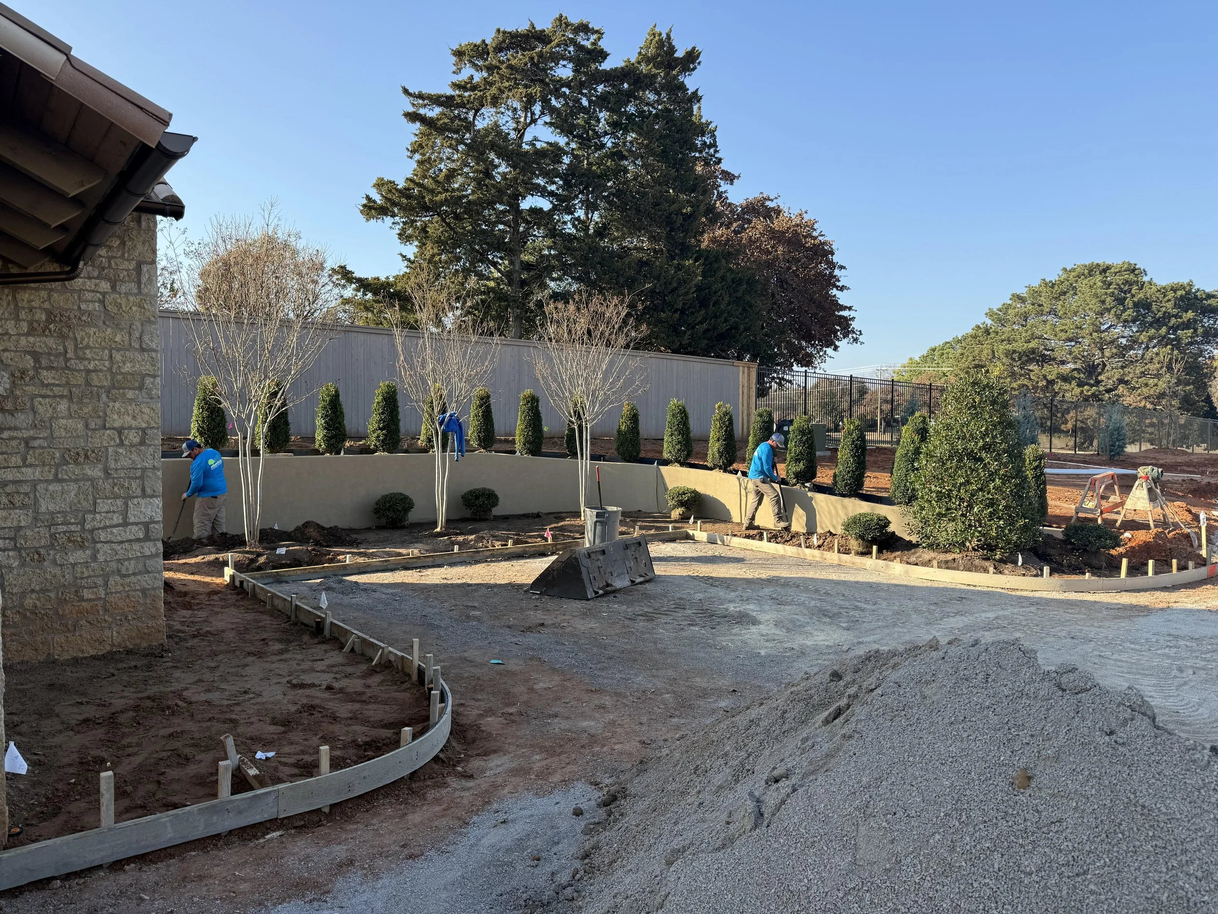 Construction workers landscaping a yard with trees, shrubs, and a curved retaining wall under a clear blue sky.
