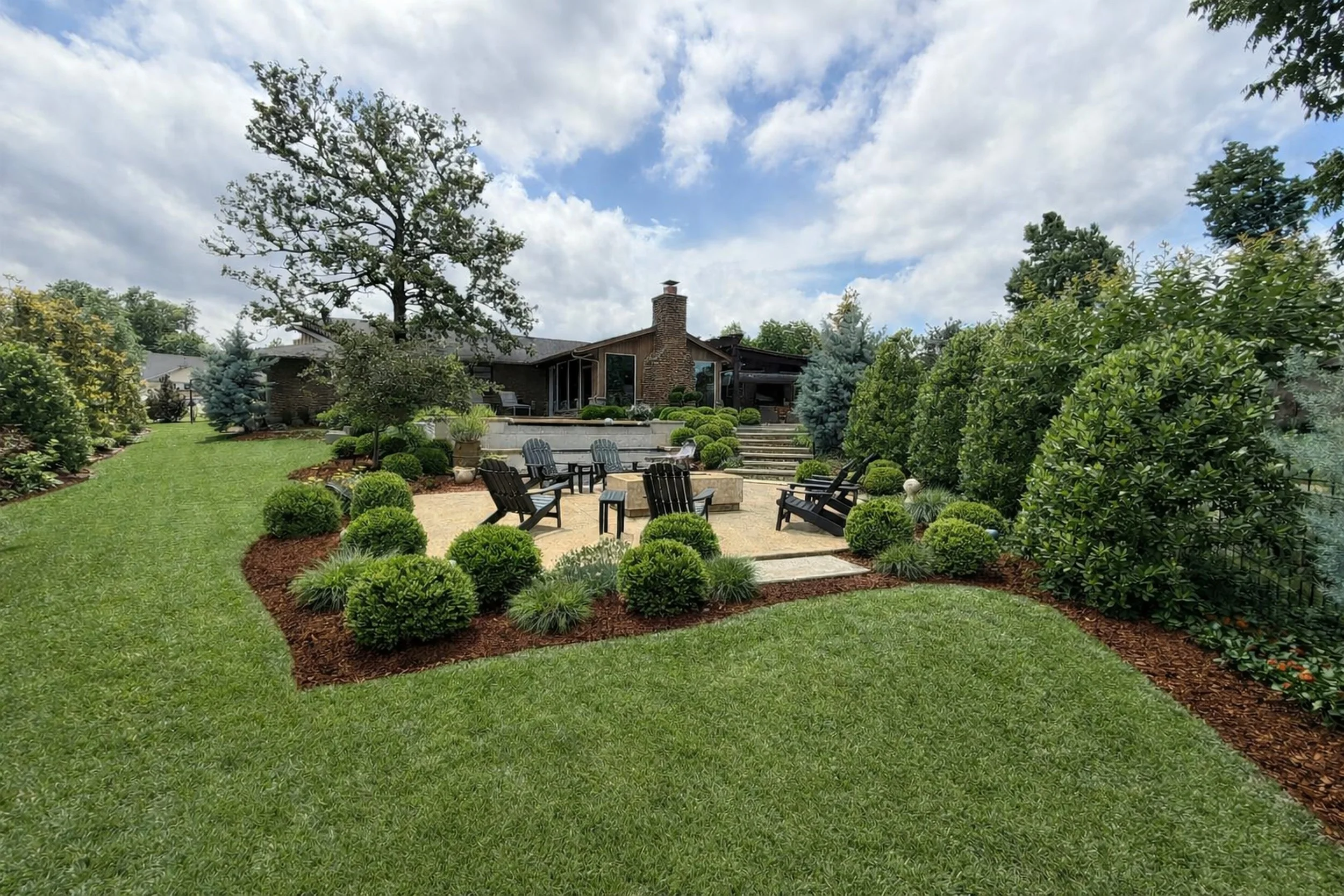 Backyard garden with neatly trimmed bushes, a grassy lawn, a stone patio with four black Adirondack chairs, and a house with a brick chimney in the background under a partly cloudy sky.