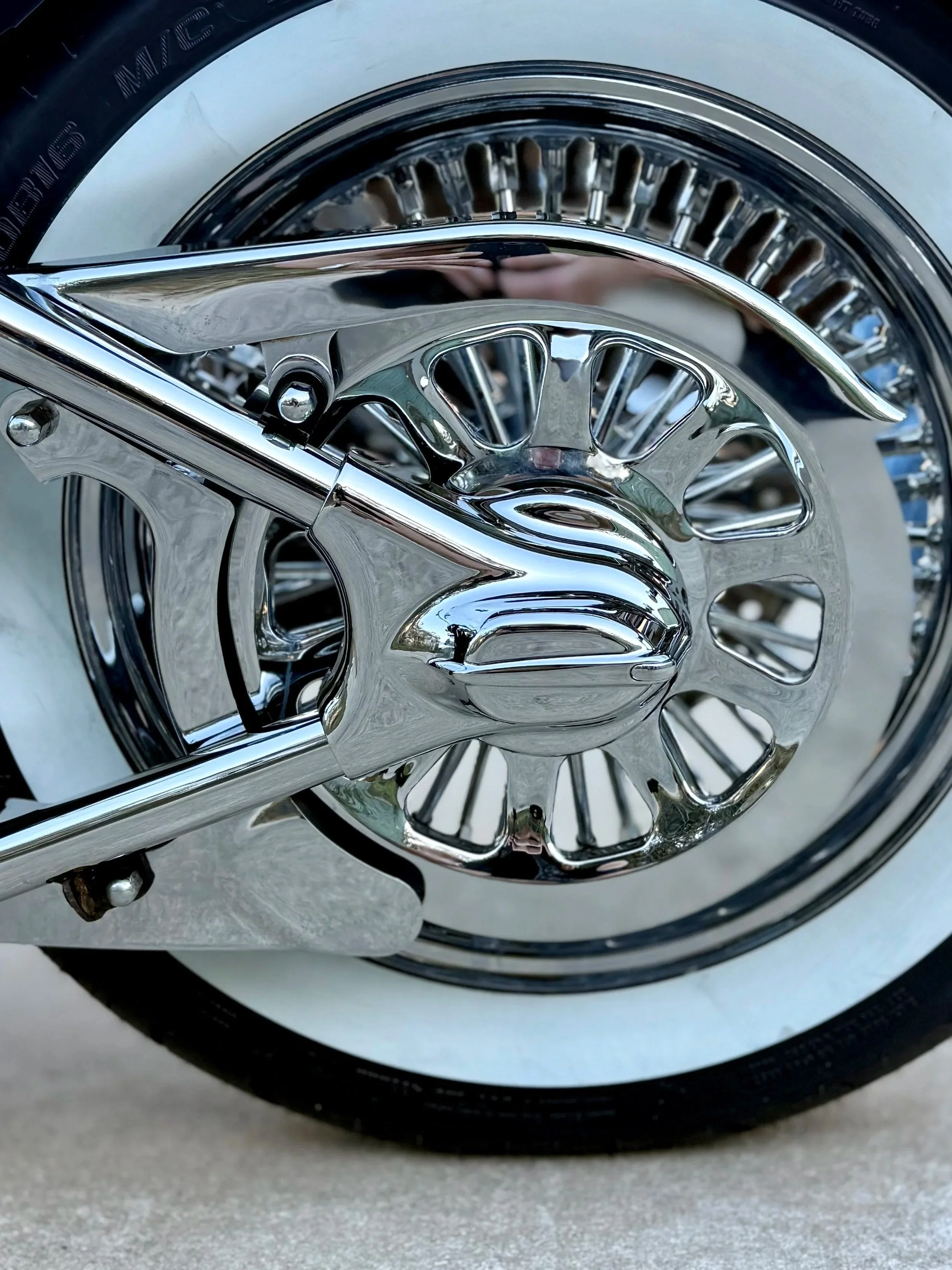 Close-up of a chrome motorcycle front wheel and hub, with whitewall tire and reflective spokes.