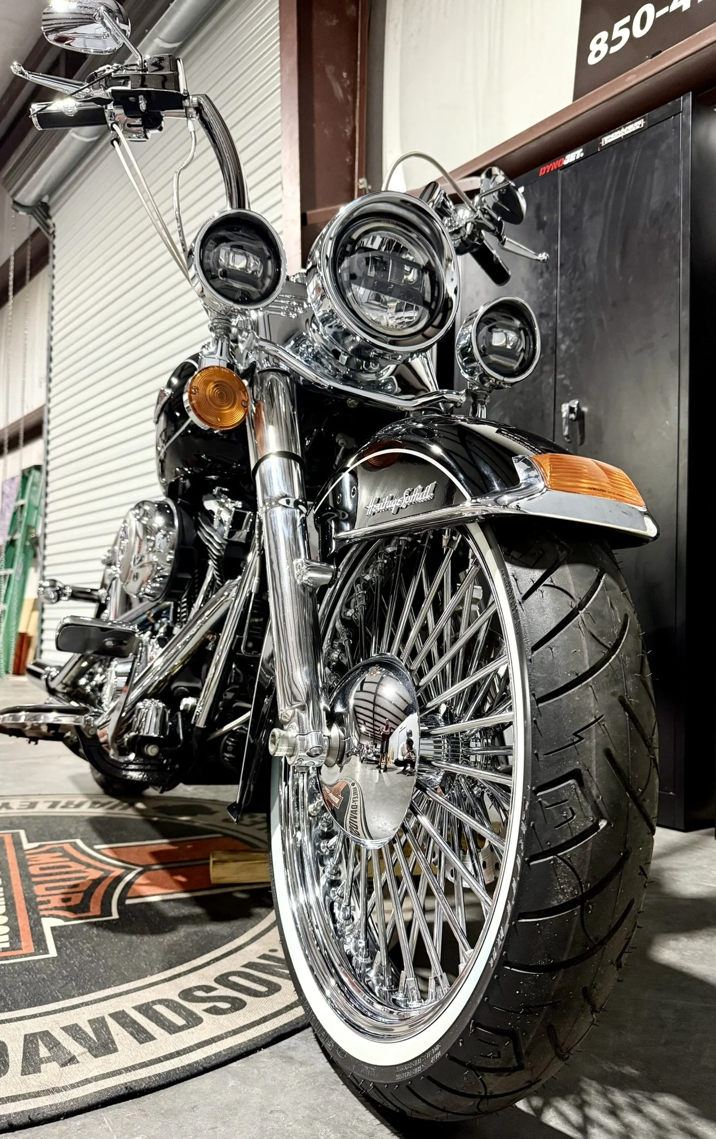 A shiny black Harley-Davidson motorcycle with chrome details, whitewall tires, and multiple headlights, parked indoors on a Harley-Davidson branded mat.