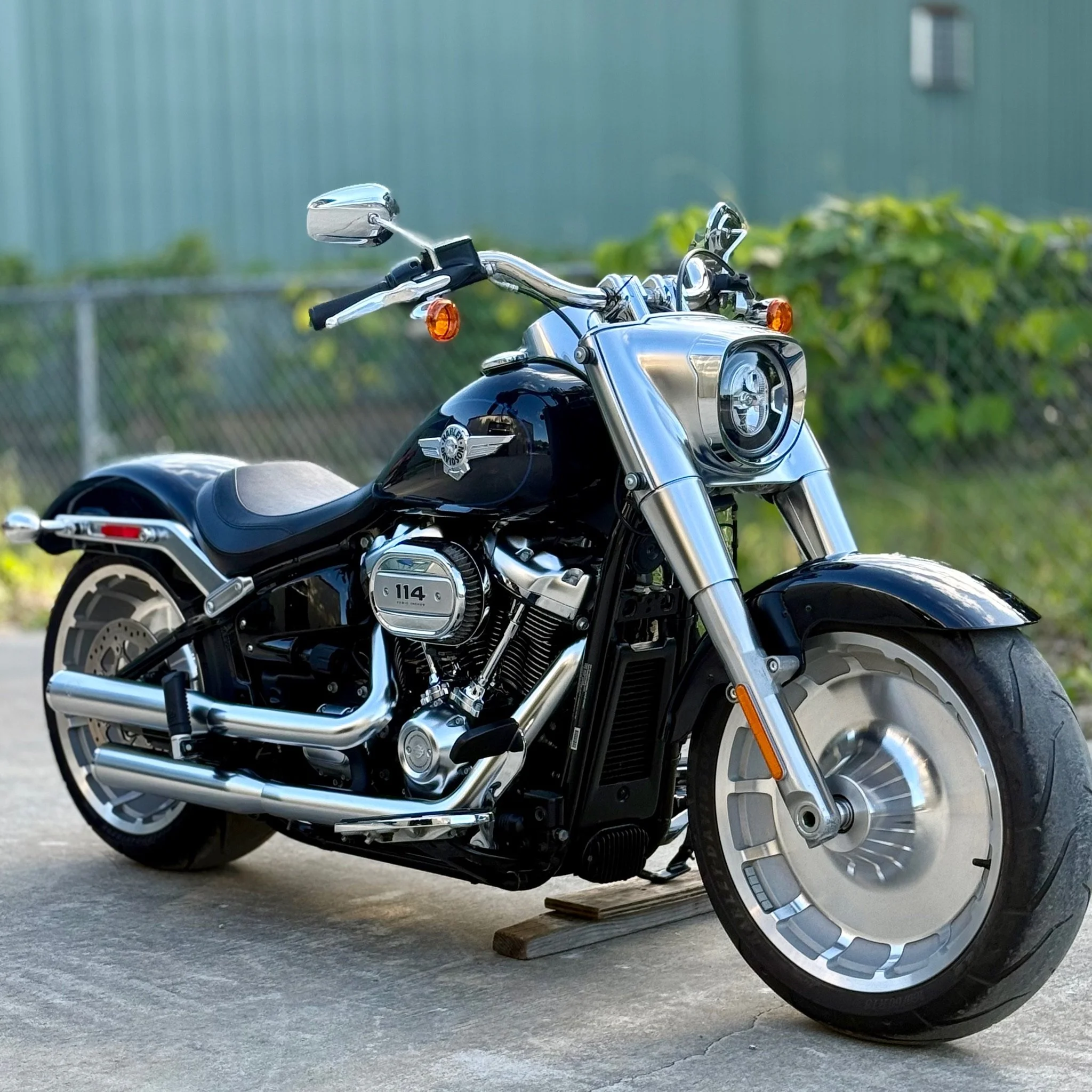 Black and silver Harley-Davidson motorcycle parked on concrete with a green fence and foliage in the background.
