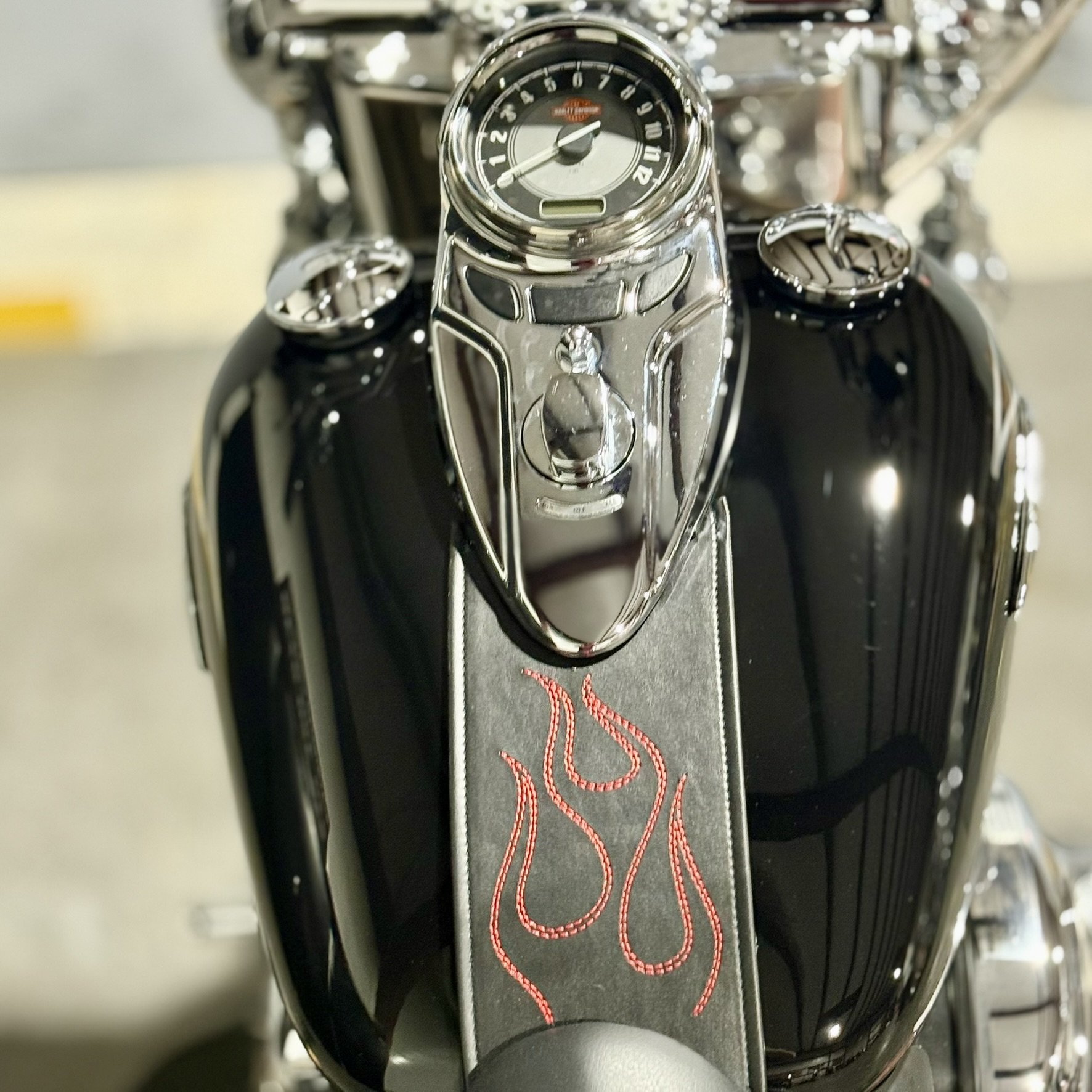 Close-up of a motorcycle's handlebar featuring a speedometer, chrome accents, and a black leather strap with red flame embroidery.