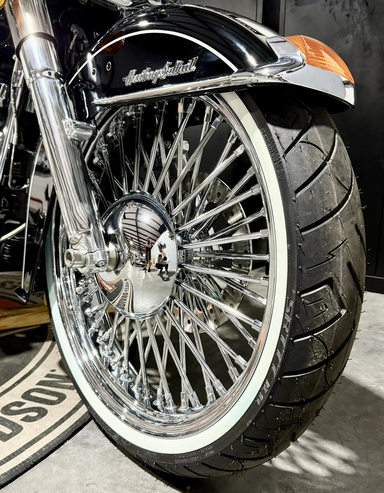 Close-up of the front wheel of a Harley-Davidson motorcycle with a chrome spoke wheel, whitewall tire, and a black front fender with Harley-Davidson logo.
