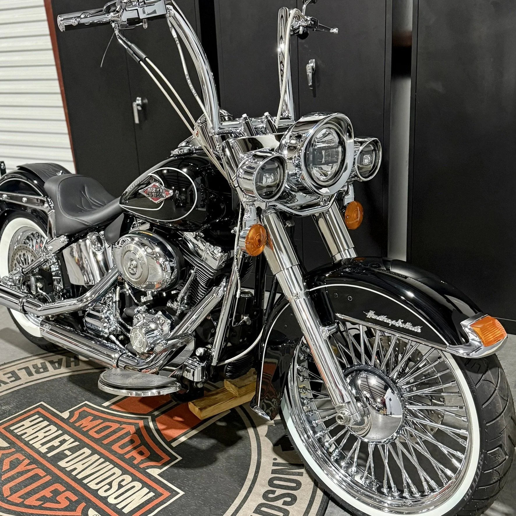A black Harley-Davidson motorcycle equipped with chrome accents, a large front wheel with multiple spokes, and a front fender that has the Harley-Davidson logo. The bike is parked on a Harley-Davidson branded mat inside a garage or showroom, with loc