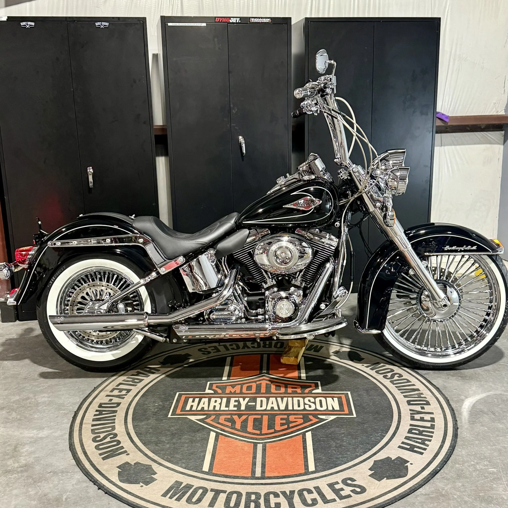 A black and chrome Harley-Davidson motorcycle parked on a circular Harley-Davidson logo mat inside a showroom, with black lockers in the background.