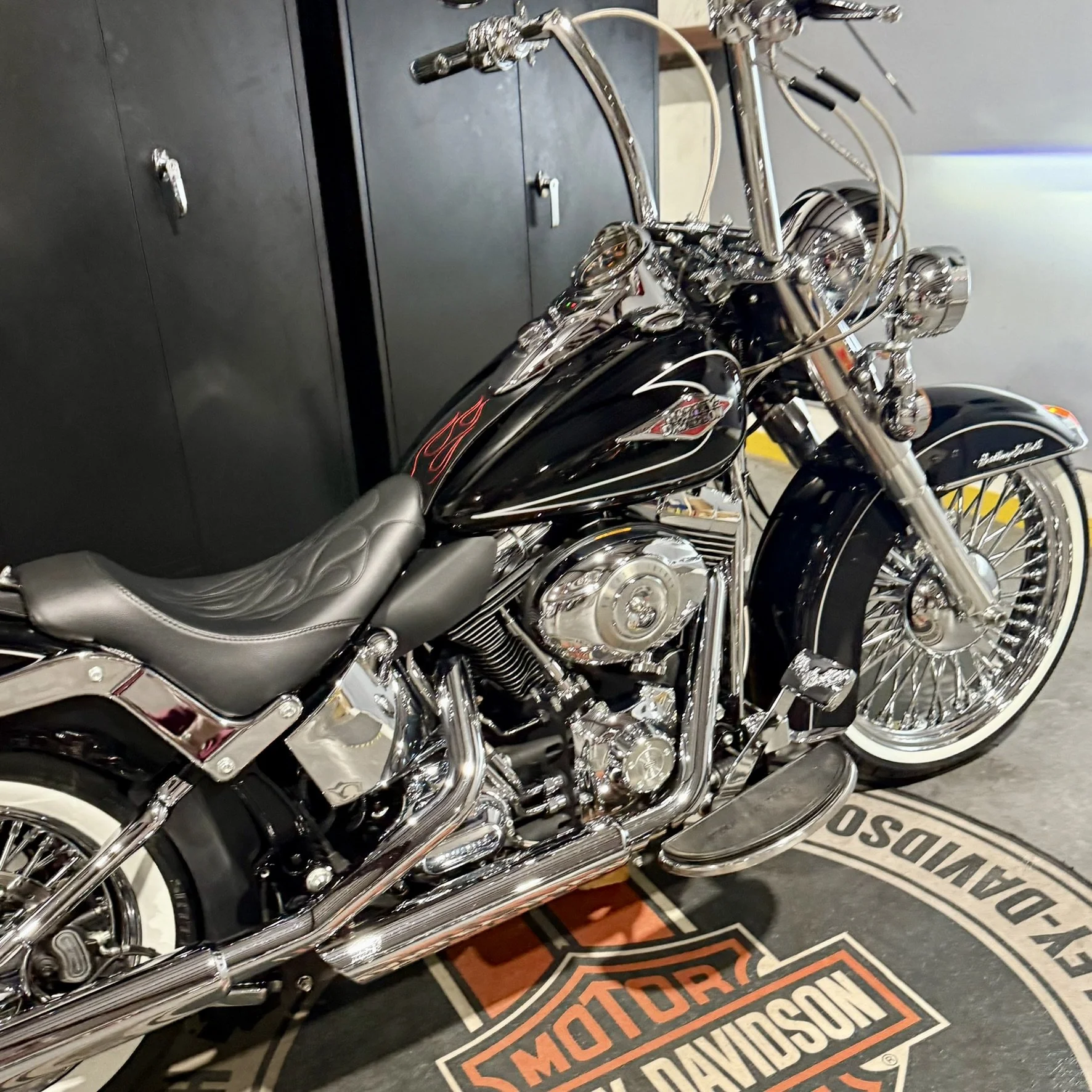 Black Harley-Davidson motorcycle with chrome accents and flame design on the tank, parked on a Harley-Davidson mat in front of black lockers.