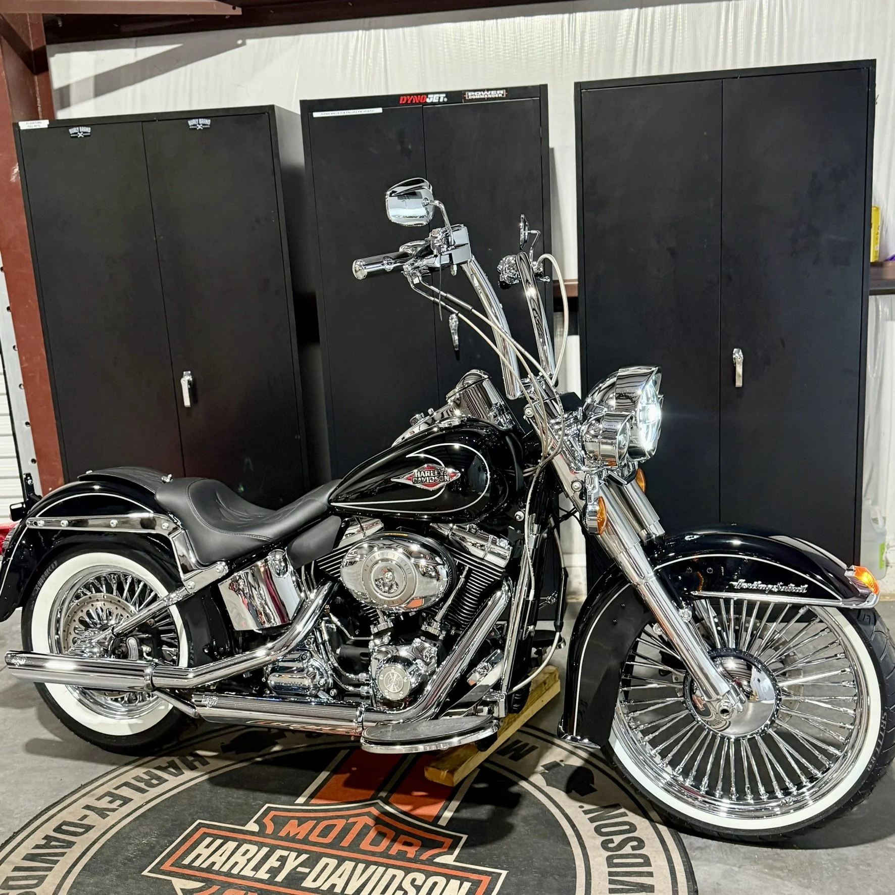 A black and chrome Harley-Davidson motorcycle with whitewall tires parked on a Harley-Davidson branded mat in front of black lockers in a garage or showroom.