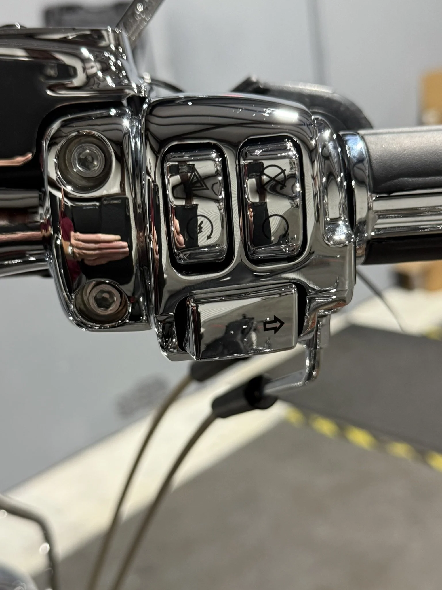 Close-up of a motorcycle handlebar control panel with chrome finish, featuring buttons for horn, turn signals, and other functions, with a reflection of the person taking the photo.