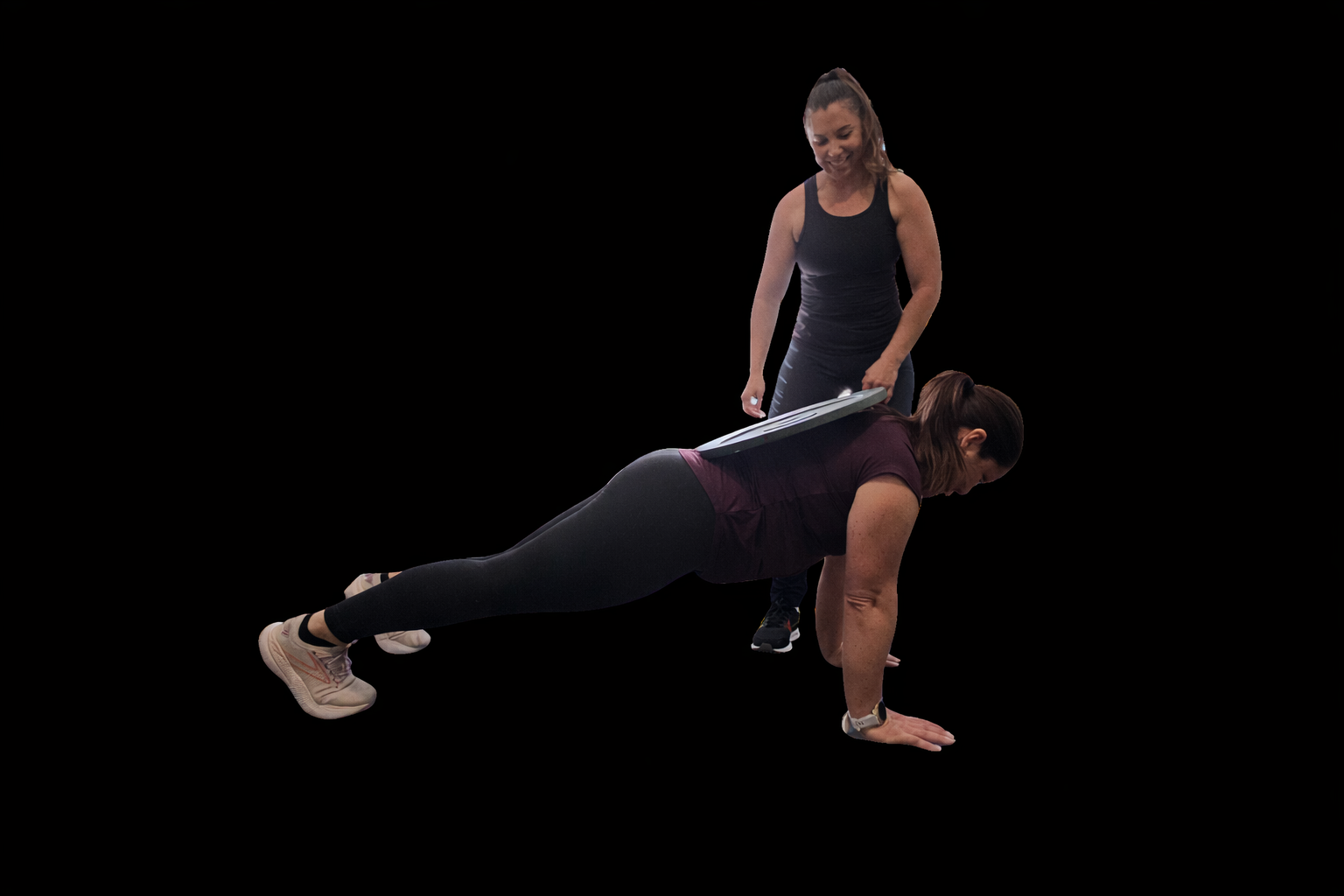 A woman performing a plank exercise on the floor while another woman assists her by placing a weight plate on her back, against a black background.
