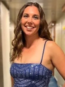 A woman with long, wavy brown hair smiling, wearing a blue lace tank top, standing indoors.