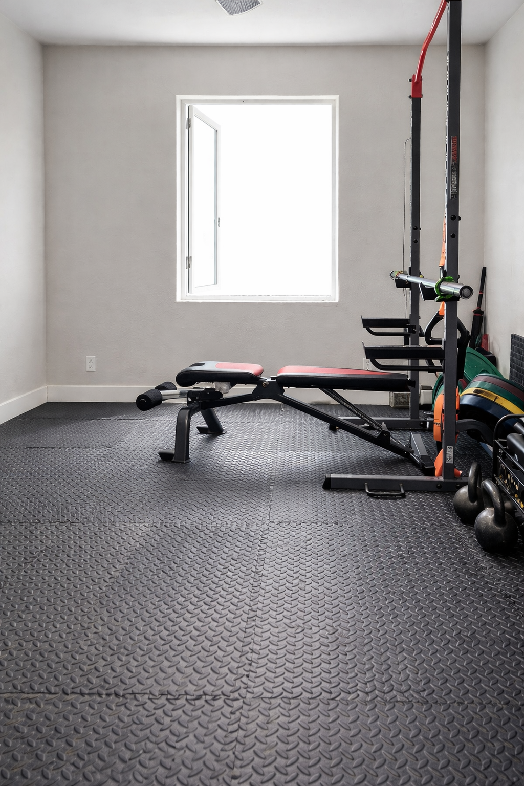 Home gym with black and red exercise bench, weight frame, and weights, illuminated by a window in a room with gray walls and textured black flooring.