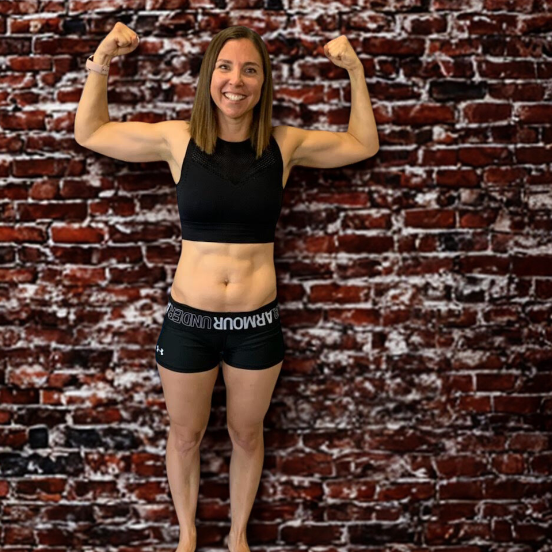 A woman smiling and flexing her arm muscles in front of a brick wall background, wearing a black sports bra and black shorts.