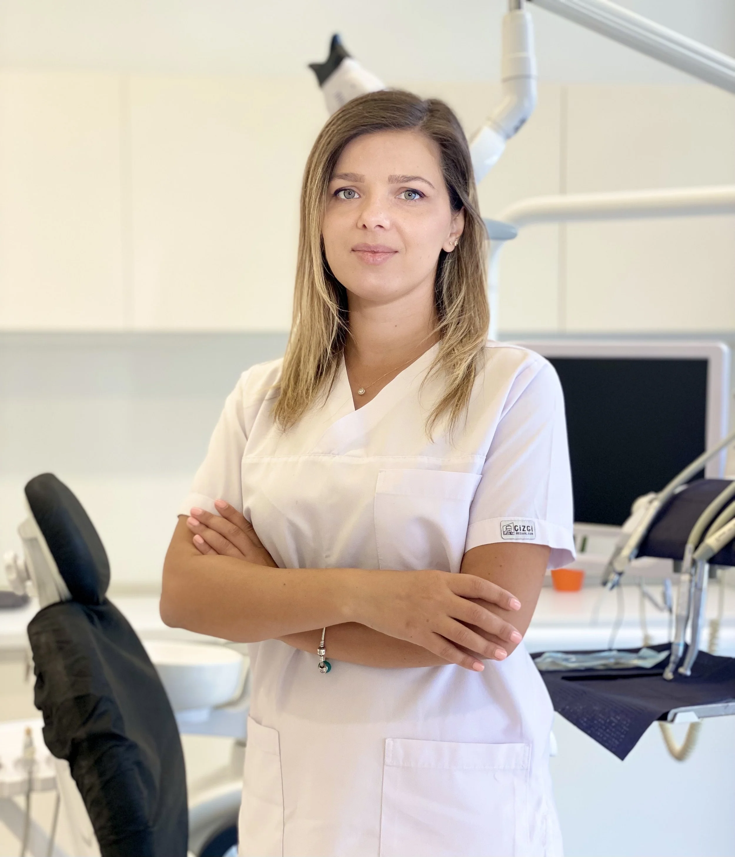 A female dentist standing in a dental clinic with arms crossed, wearing a white dental coat, in front of dental equipment and office supplies.