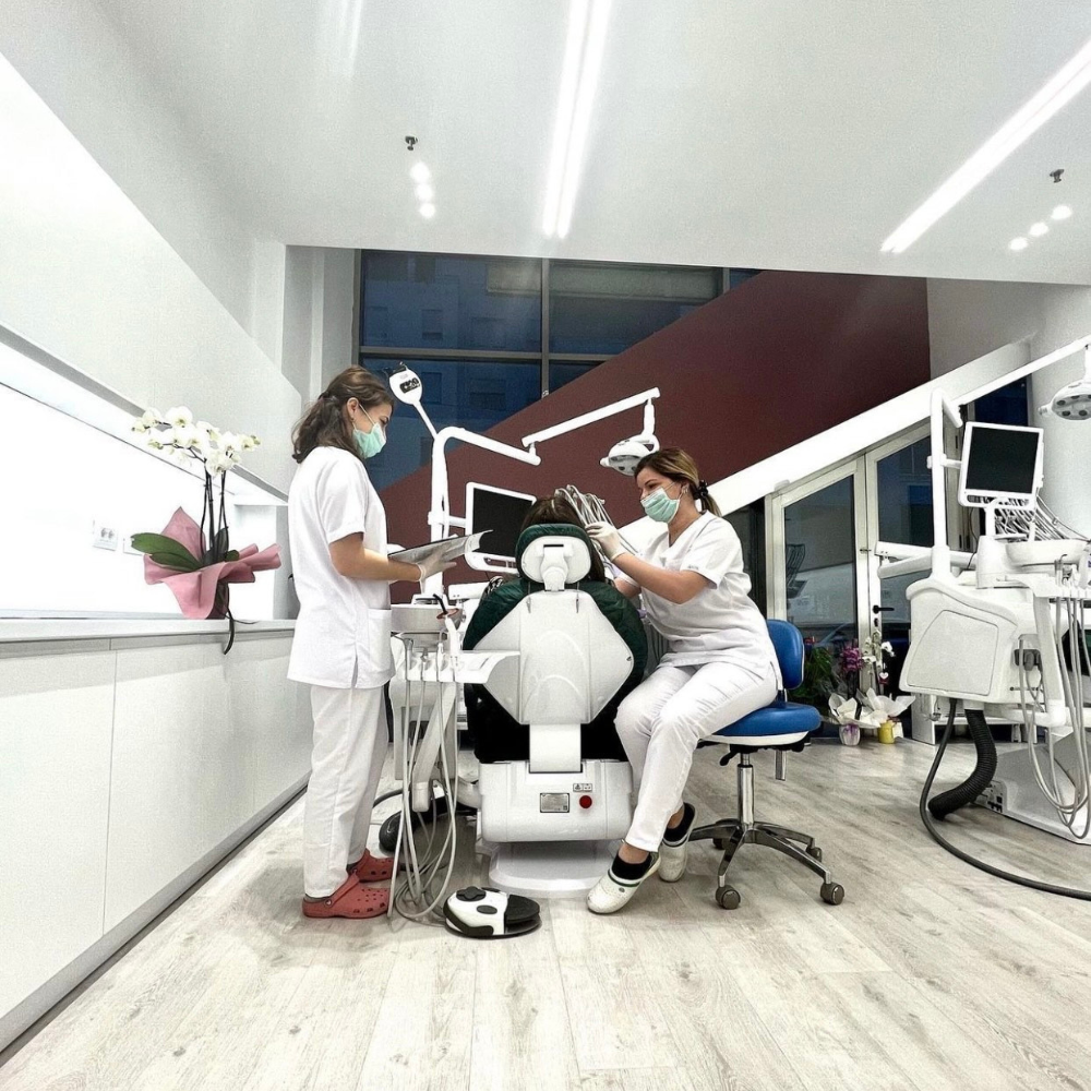 Dental clinic staff attending to a patient in a dental chair, with medical equipment and monitors around.