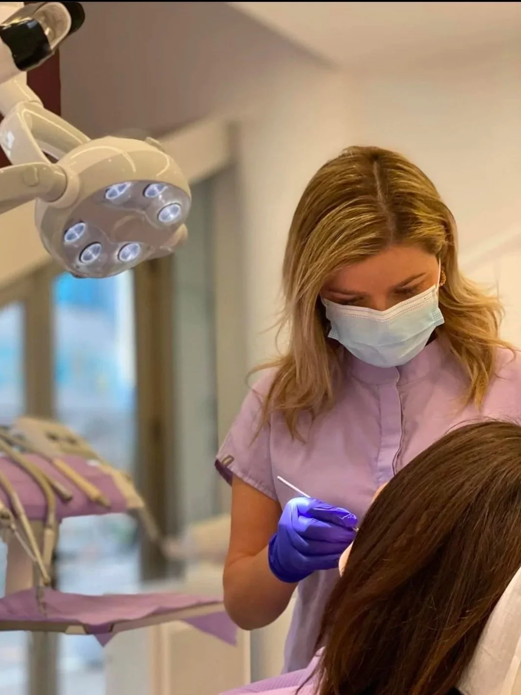 A female dental professional wearing a mask and gloves performing a dental procedure on a patient, with dental lights overhead.
