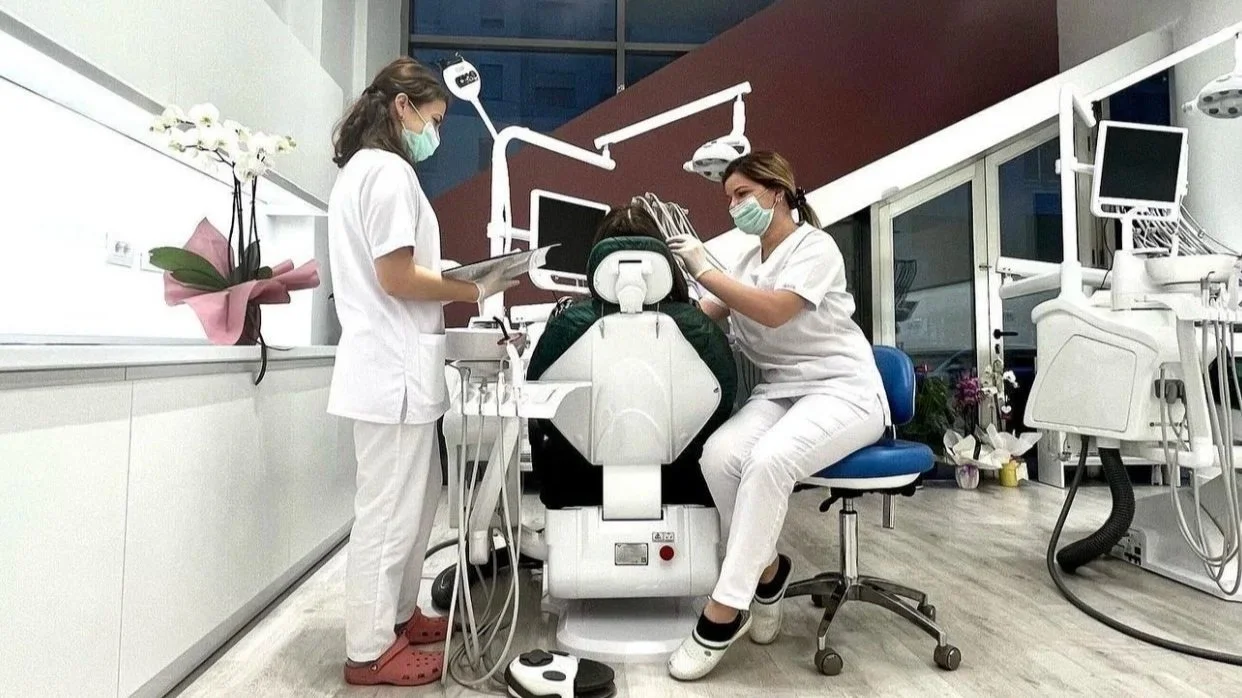 Two dental professionals attending to a patient in a dental clinic, with dental equipment and monitors around.