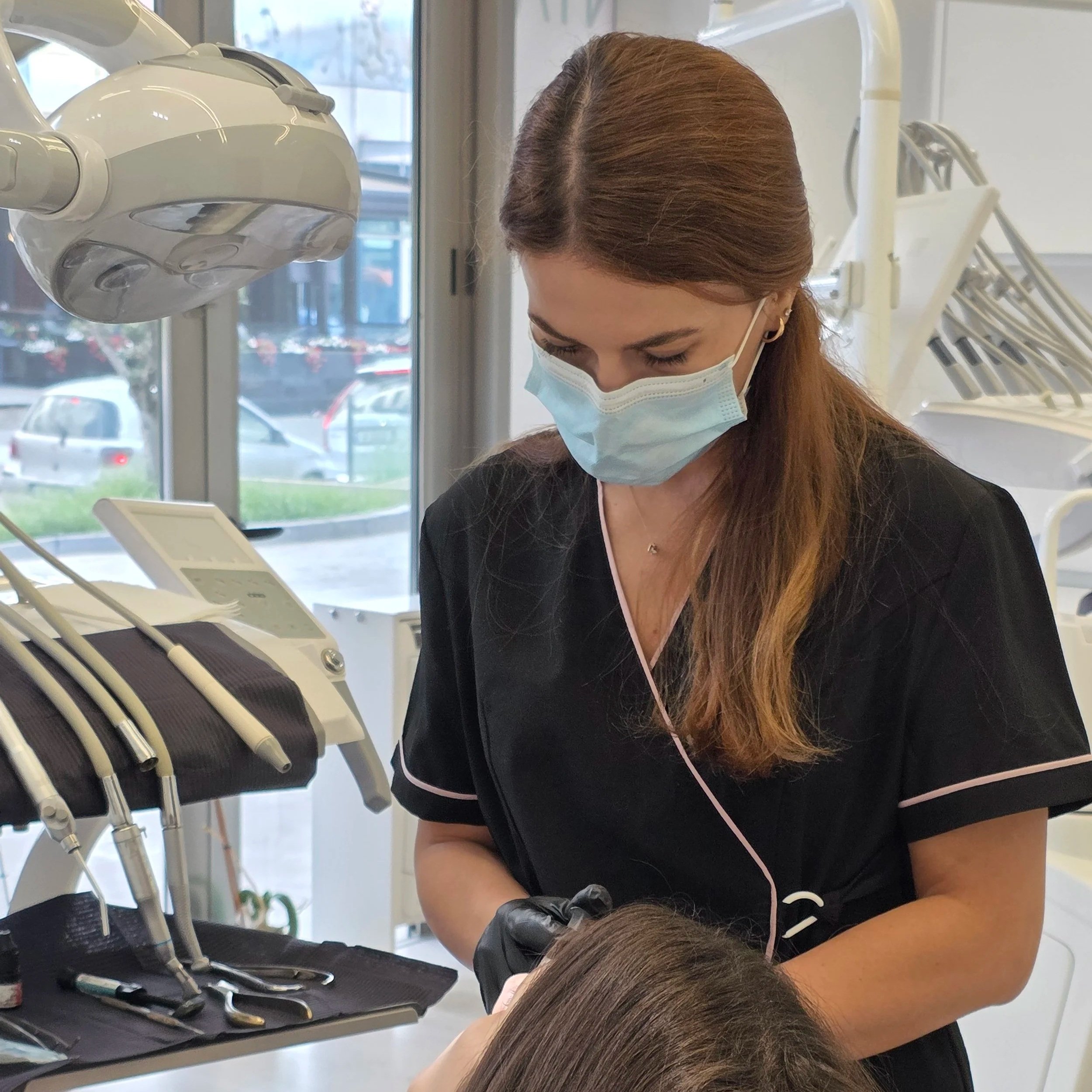 A female dental professional wearing a face mask and gloves working on a patient in a dental office with dental tools and equipment visible.