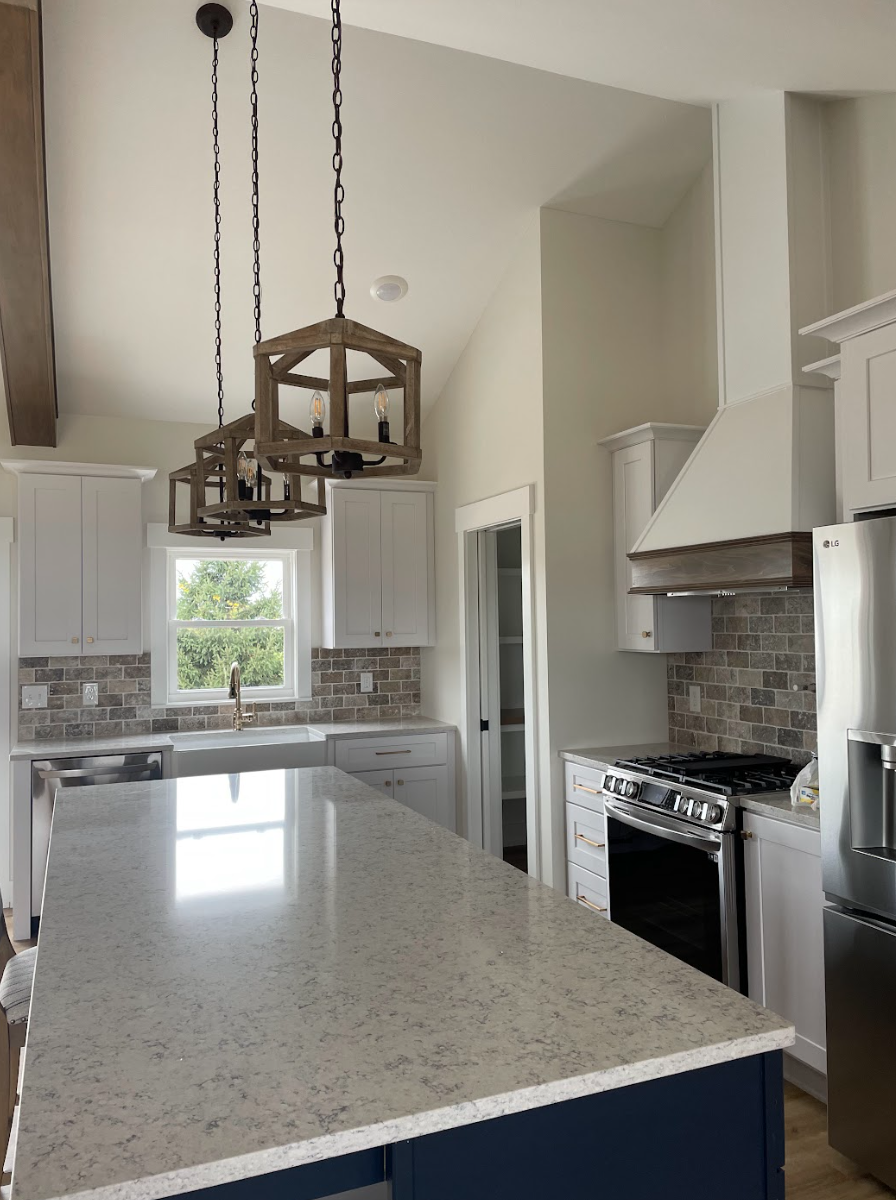 Modern kitchen with white cabinets, brick backsplash, granite countertop island, stainless steel appliances, window with greenery outside, and wooden pendant light fixtures.