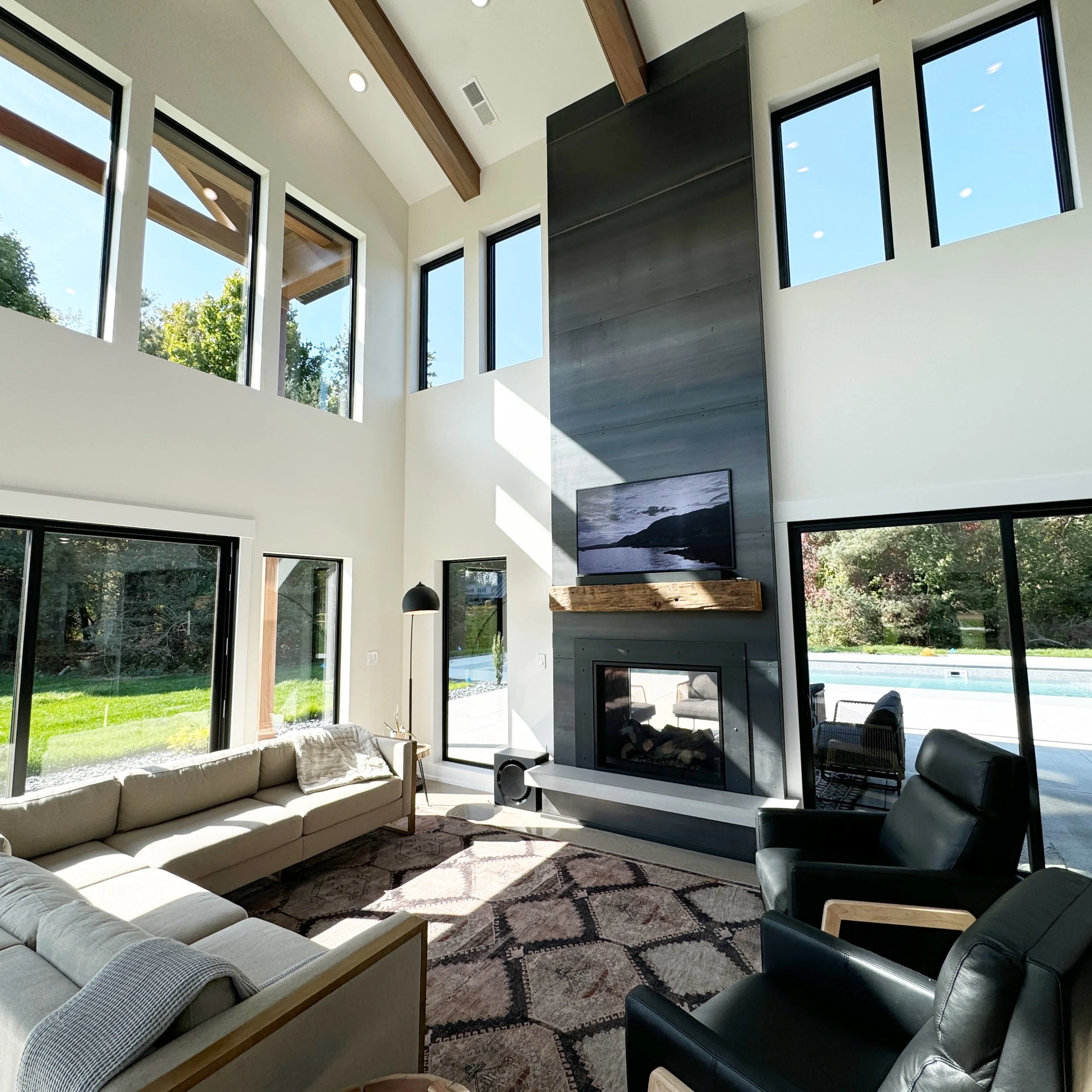 Modern living room with high ceilings, large windows, a fireplace with a mounted television, and a mix of beige and black furniture.