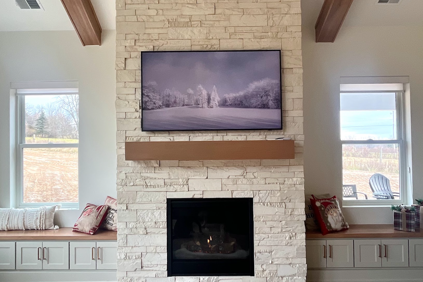 Living room with a stone fireplace, television mounted above, two windows with window seats, holiday pillows and gifts on right side window seat.