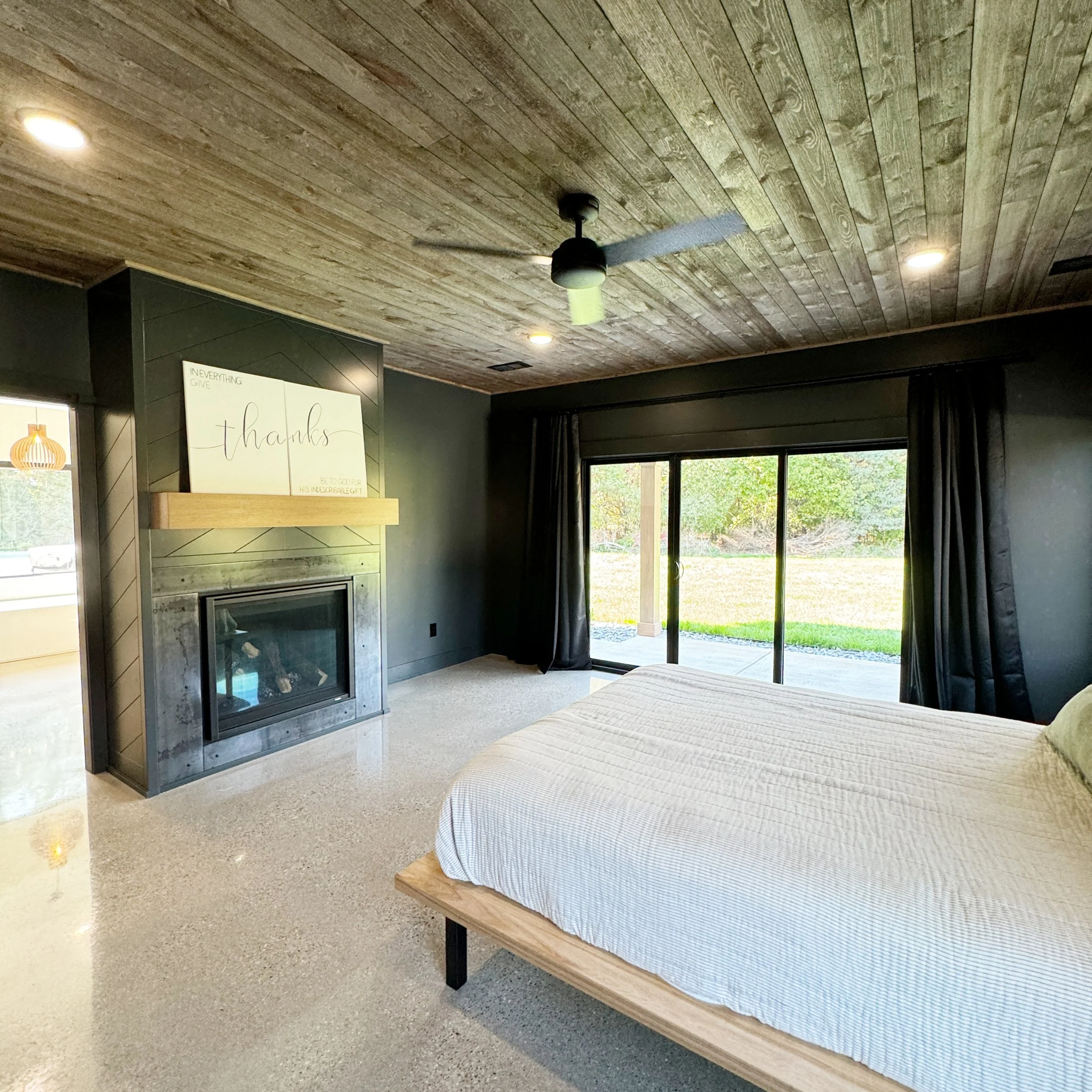 A bedroom with a modern fireplace, a wooden ceiling fan, a large sliding glass door with dark curtains, a bed with a light-colored quilt, and a view of a backyard with trees.