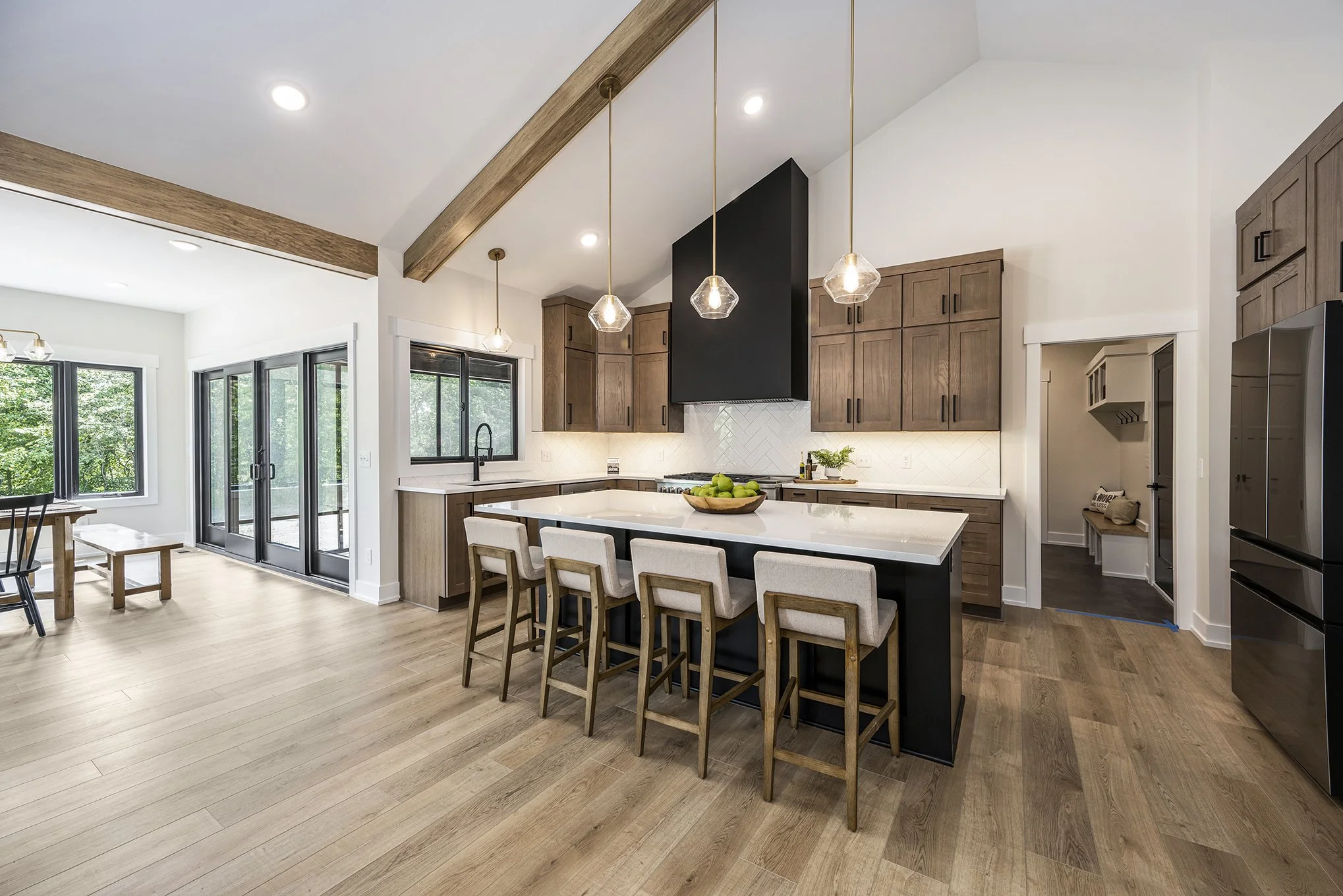 Modern kitchen with wooden cabinets, white countertop, island with beige chairs, black pendant lights, black range hood, and large windows.