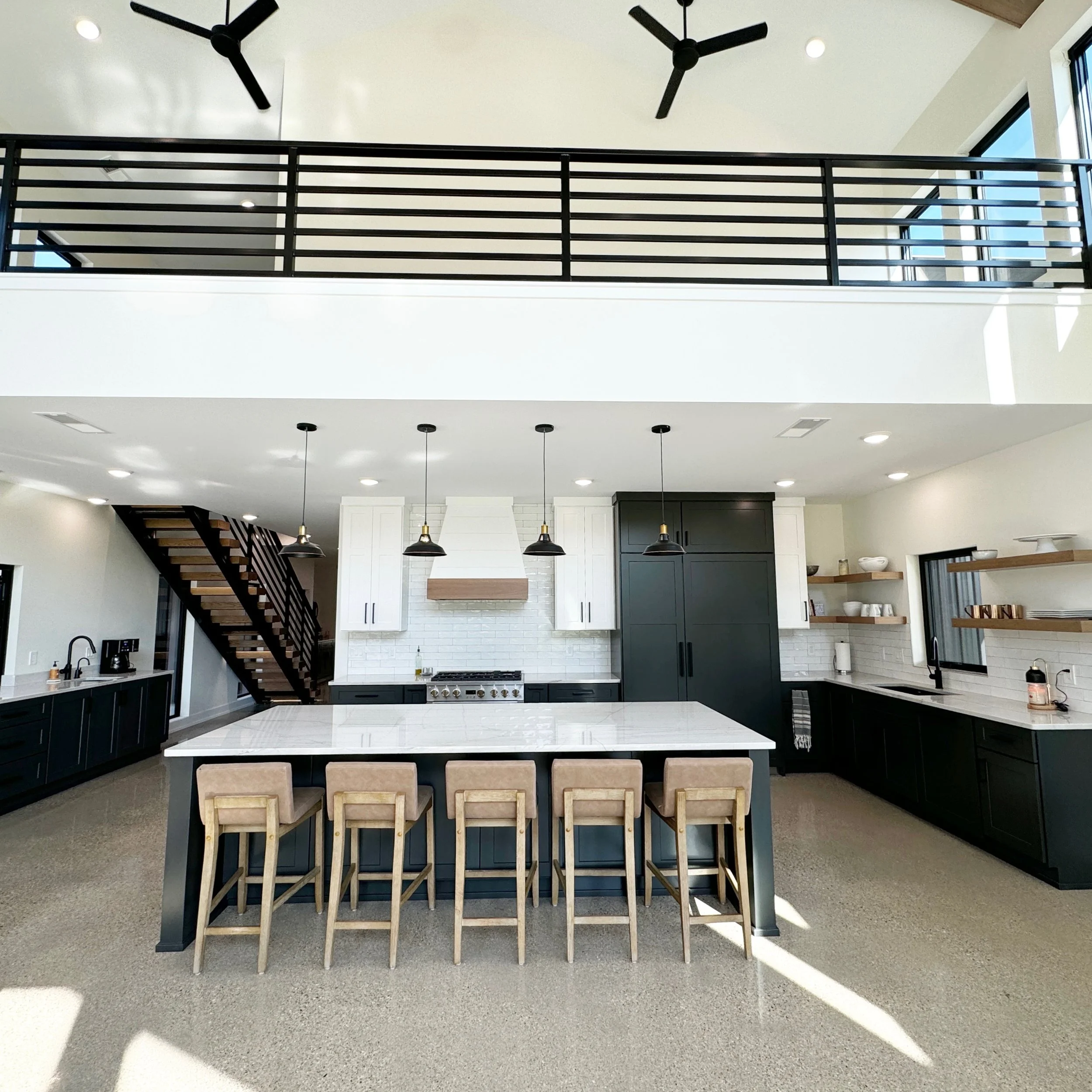 Modern kitchen with black cabinets, white countertops, a large central island with six beige upholstered chairs, pendant lights, a white range hood, open shelving with dishes, and a staircase leading upstairs.