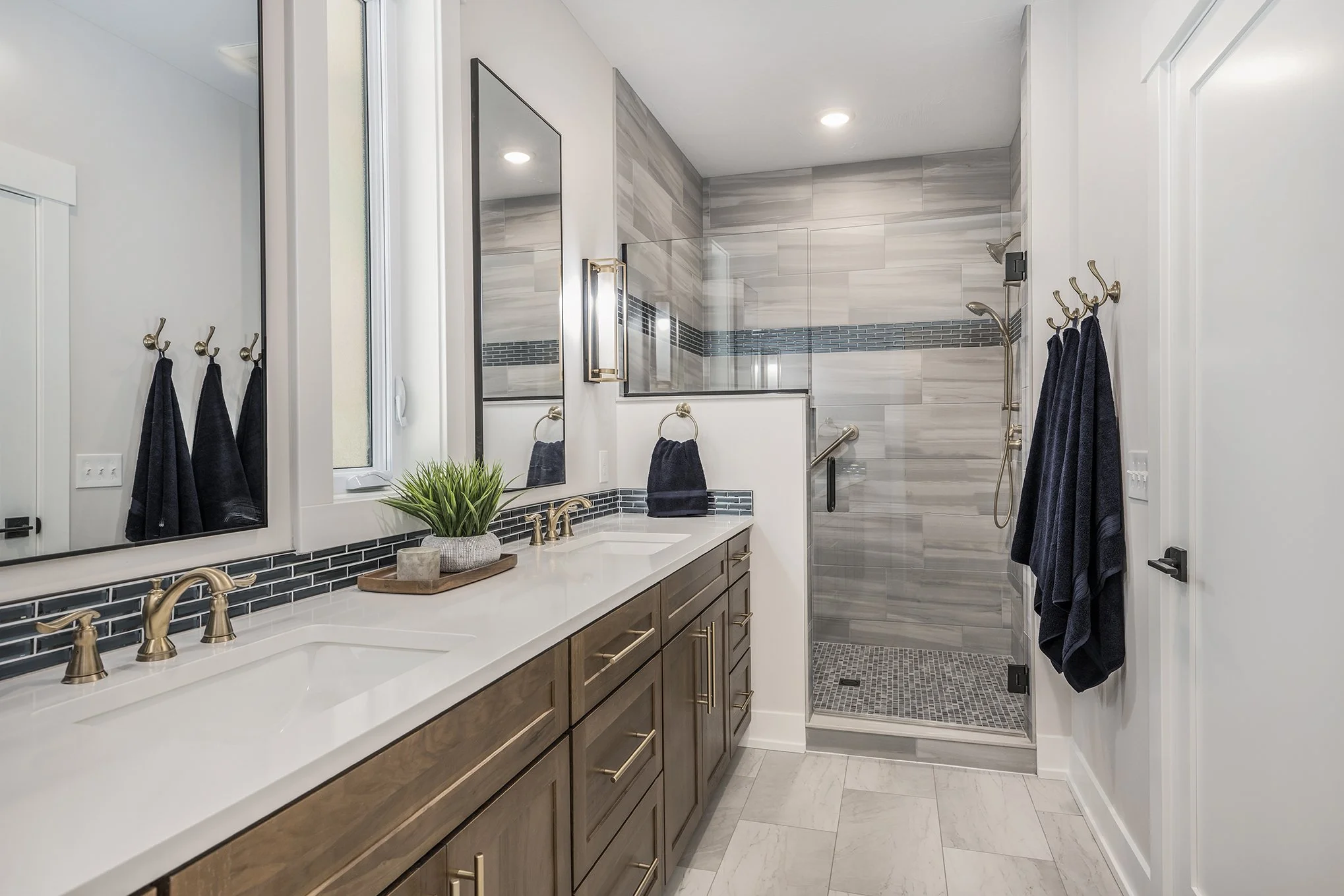 Modern bathroom with double vanity, wooden cabinets, large mirror, gold fixtures, walk-in glass shower with gray tiles, navy towels, and a small potted plant