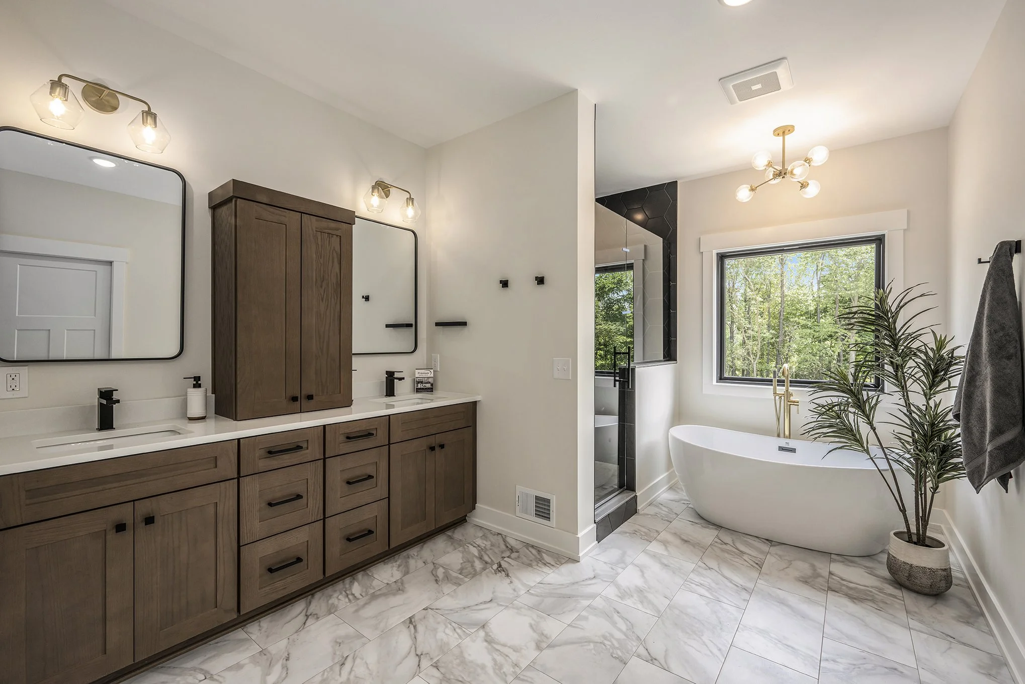 Modern bathroom with wooden vanity, double mirrors, walk-in shower, freestanding bathtub, window overlooking trees, and a potted plant.