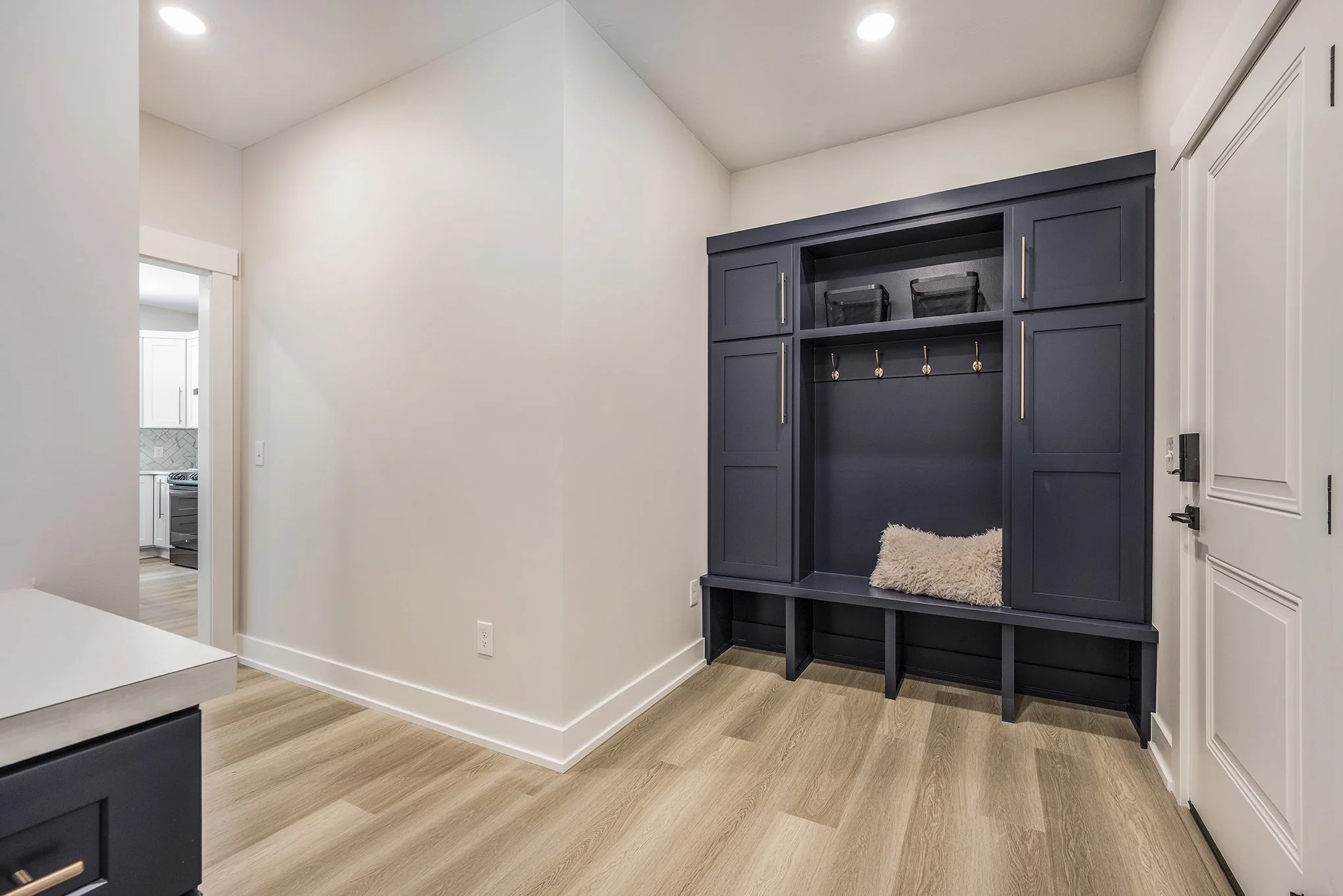 Interior view of a modern home with a built-in navy blue & black mudroom locker with hooks, baskets, and a fluffy pillow, white walls, light wood flooring, and a view into the kitchen.