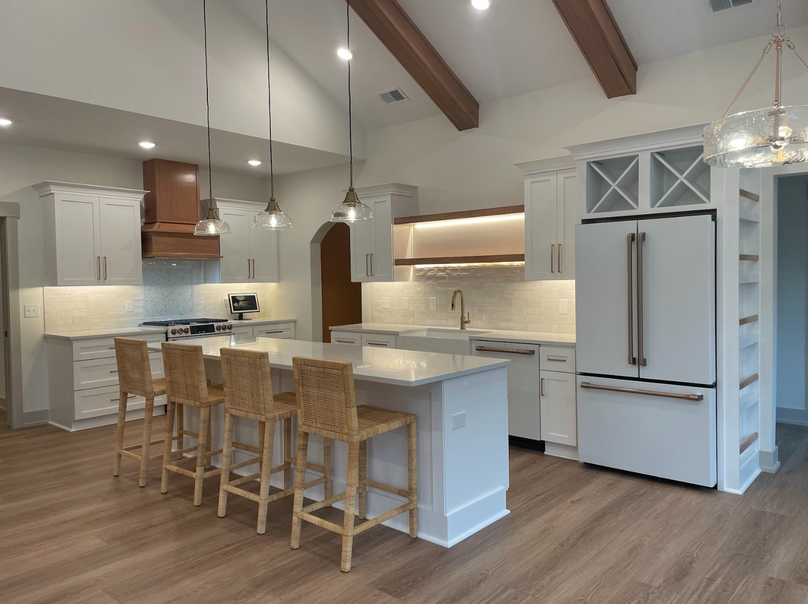 Modern white kitchen with island, four wicker barstools, recessed lighting, pendant lights, wooden beams, white cabinets, stainless steel appliances, hardwood floor.
