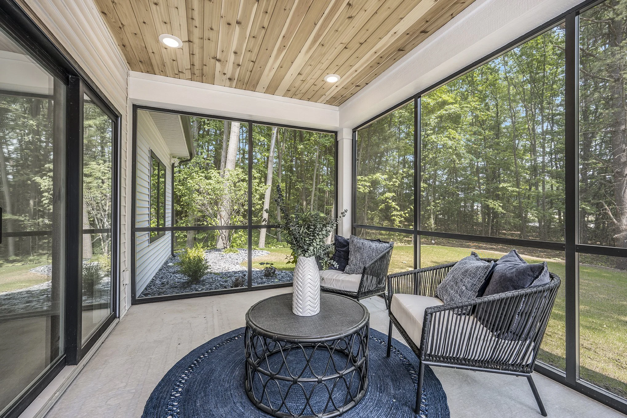 Sunroom with black-framed glass walls, beige concrete floor, and wood-paneled ceiling. Contains two black wicker chairs with cushions, a round black coffee table with a white vase of greenery, and a navy blue rug. Outside is a wooded yard with trees 