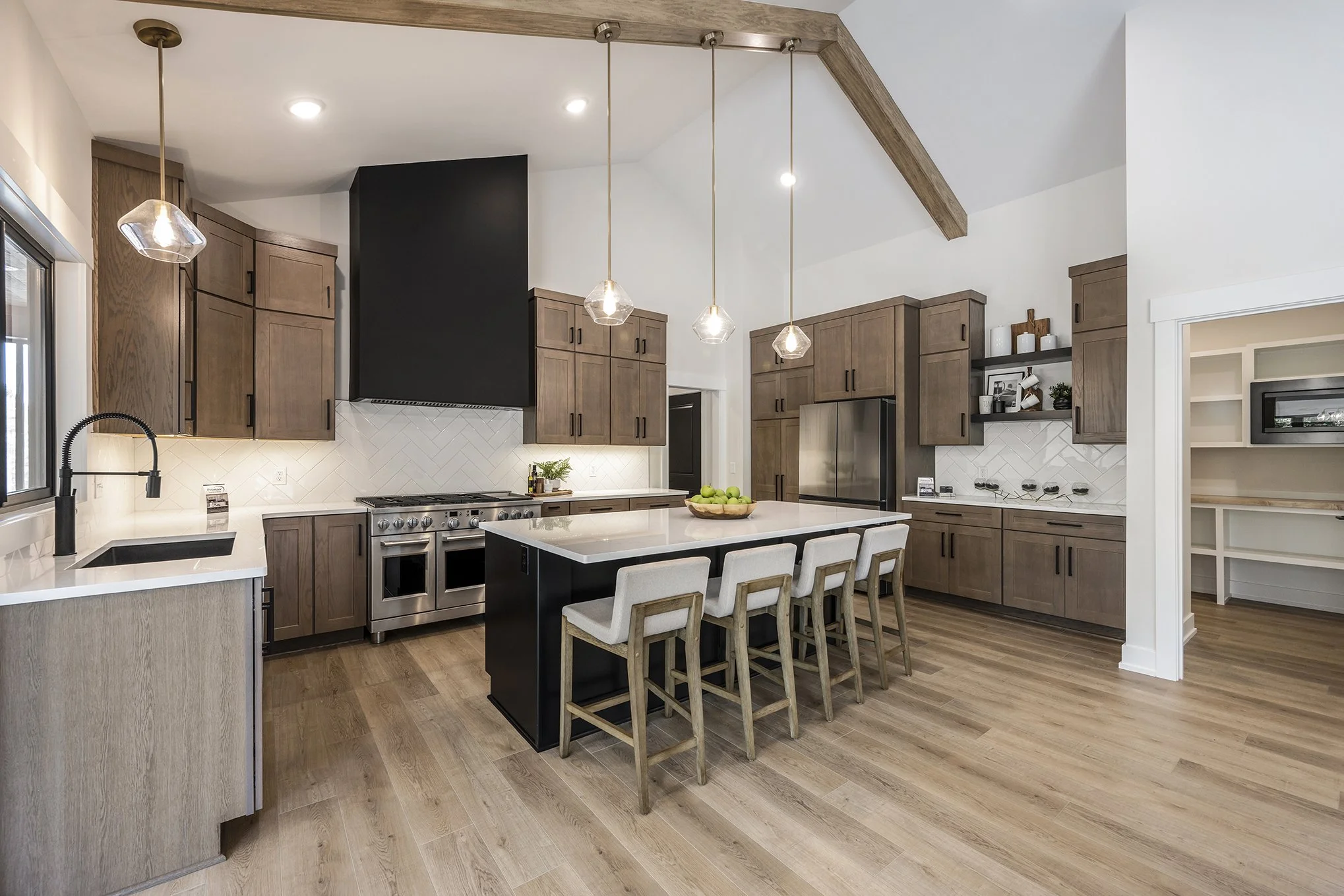 Modern kitchen with wooden cabinets, a black range hood, and a kitchen island with four chairs. Three pendant lights hang above the island. The floor is wooden, and there are white tiled backsplashes.