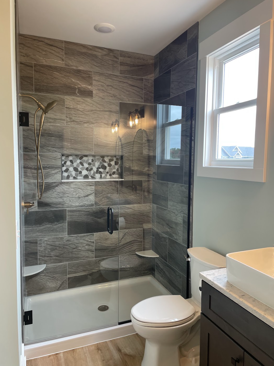A modern bathroom featuring a glass-enclosed shower with marble-like tile walls, a built-in shower niche with pebble tile accents, and a rain showerhead. There is a small window on the right allowing natural light, and a dark-colored vanity with a ve