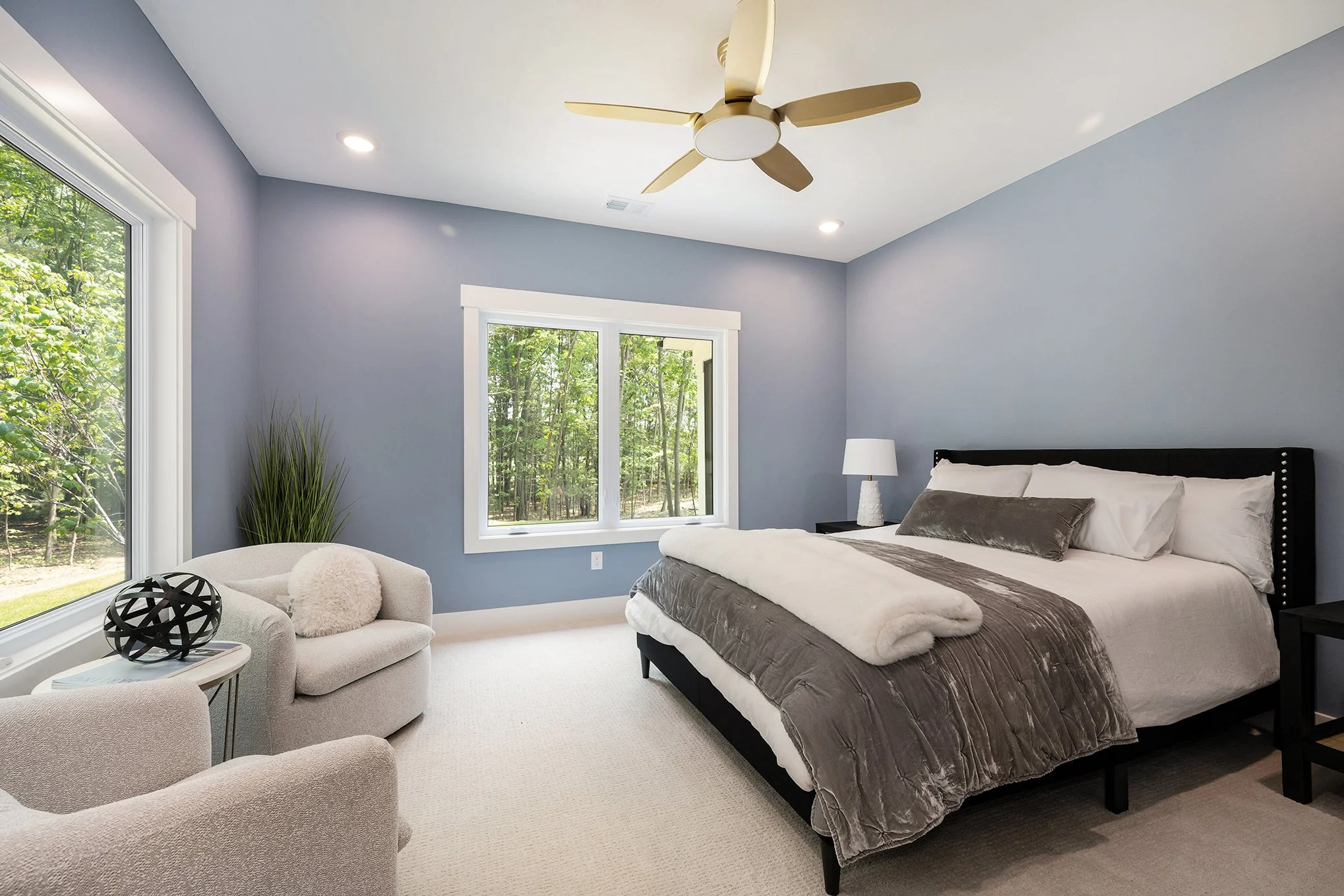 Modern bedroom with light purple walls, large windows showing a wooded outdoor scene, a white bed with gray and white bedding, a black headboard, a nightstand with a lamp, and a seating area with two chairs, an armrest, and decorative pillows.
