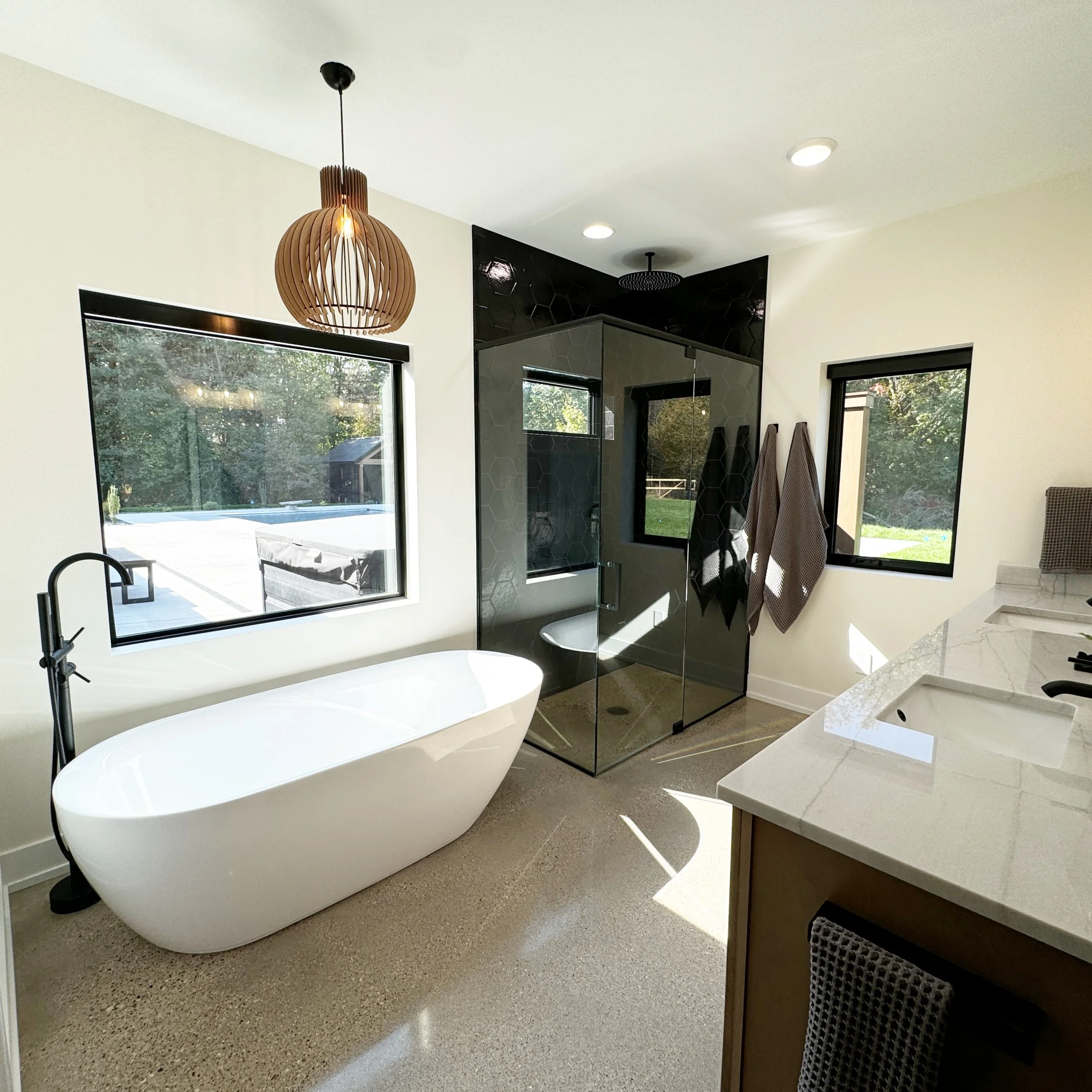 Modern bathroom with a standalone white bathtub, black fixtures, a glass shower enclosure, two black-framed windows, a marble countertop with two sinks, and towels hanging on the wall.