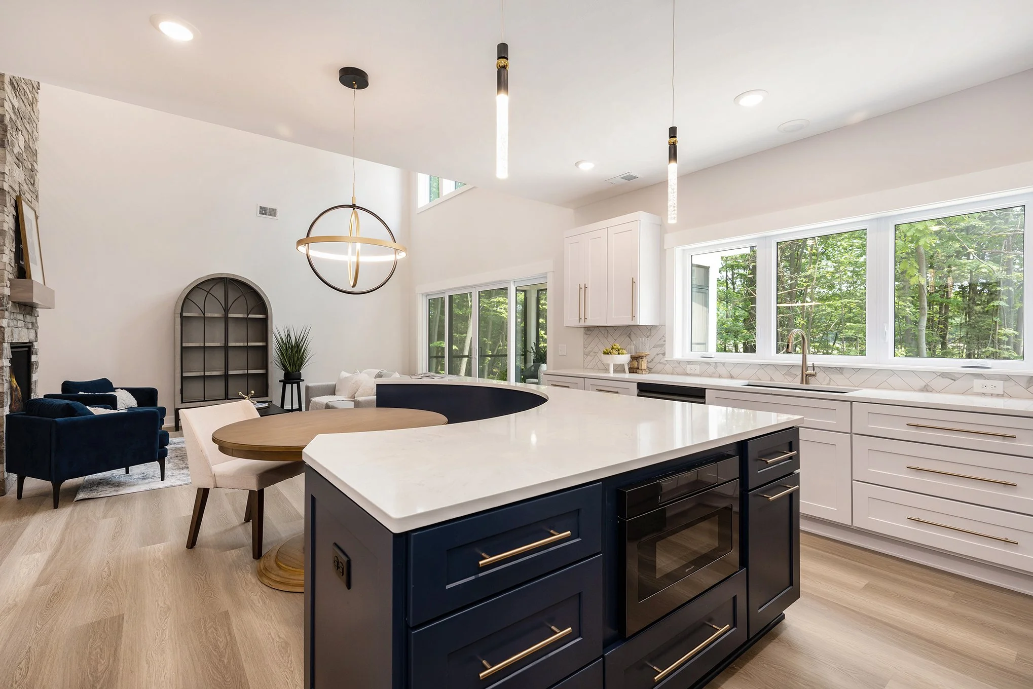 Modern open-concept kitchen with white cabinets, large windows with a view of greenery, a center island with navy blue cabinetry, a built-in oven, and a dining area with a round wooden table and beige chairs. In the background, a living area with dar