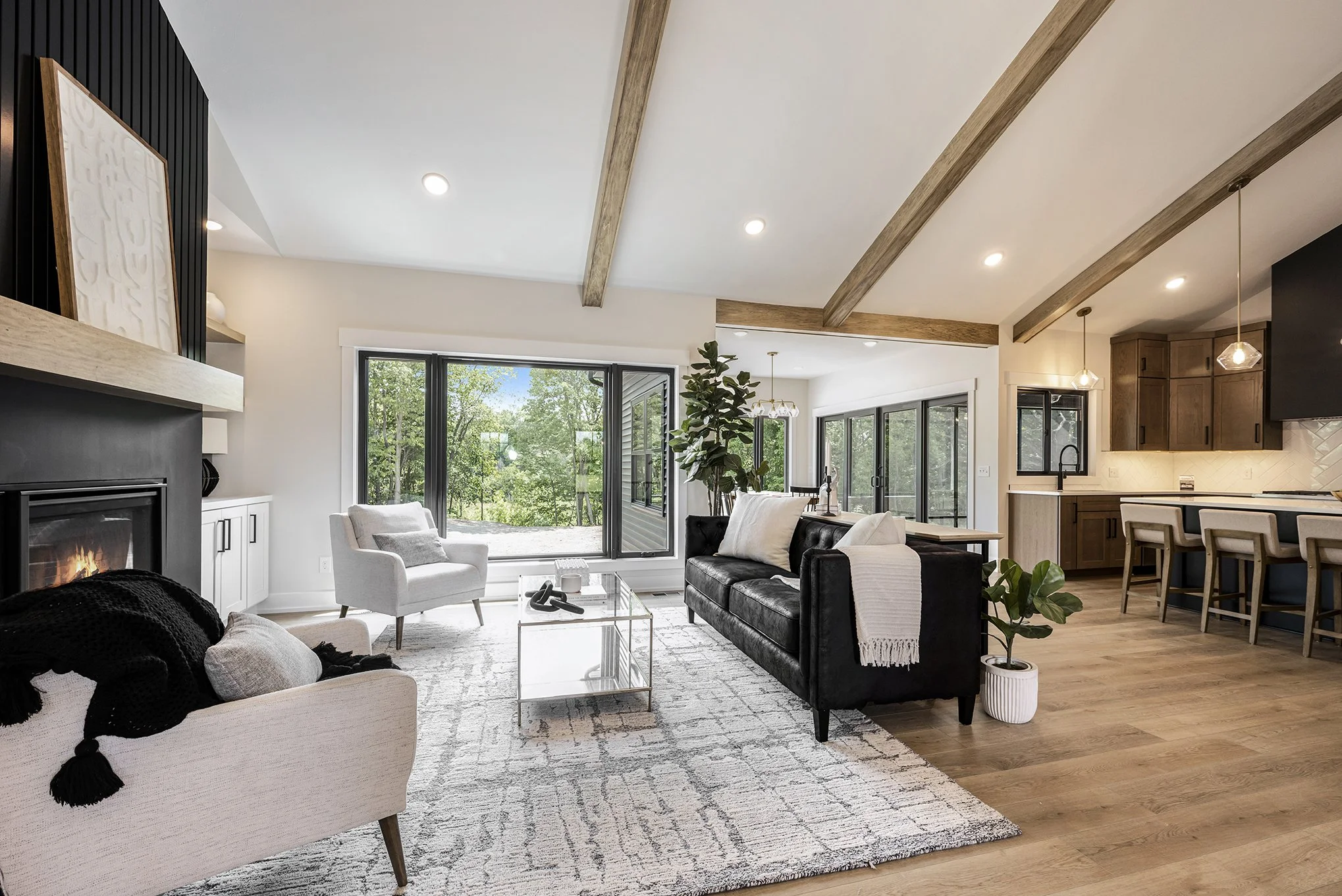 Modern open-concept living room with large window, white walls, wood beams, and hardwood floors. Contains white and black sofas, a glass coffee table, a fireplace, a large potted plant, and a view of greenery outside.