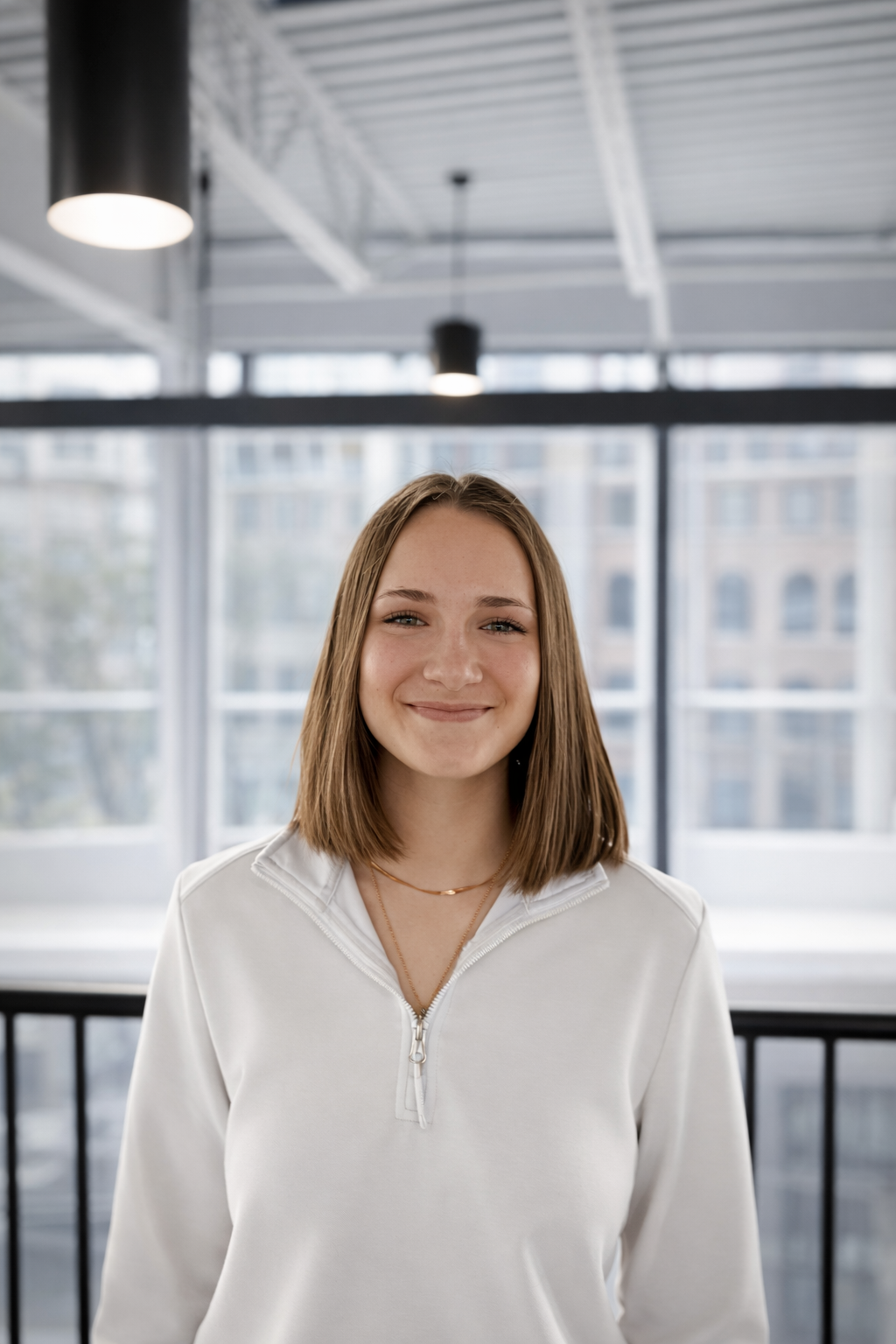A young woman with shoulder-length light brown hair, smiling, wearing a white zip-up top, in a modern indoor setting with large windows and ceiling lights.