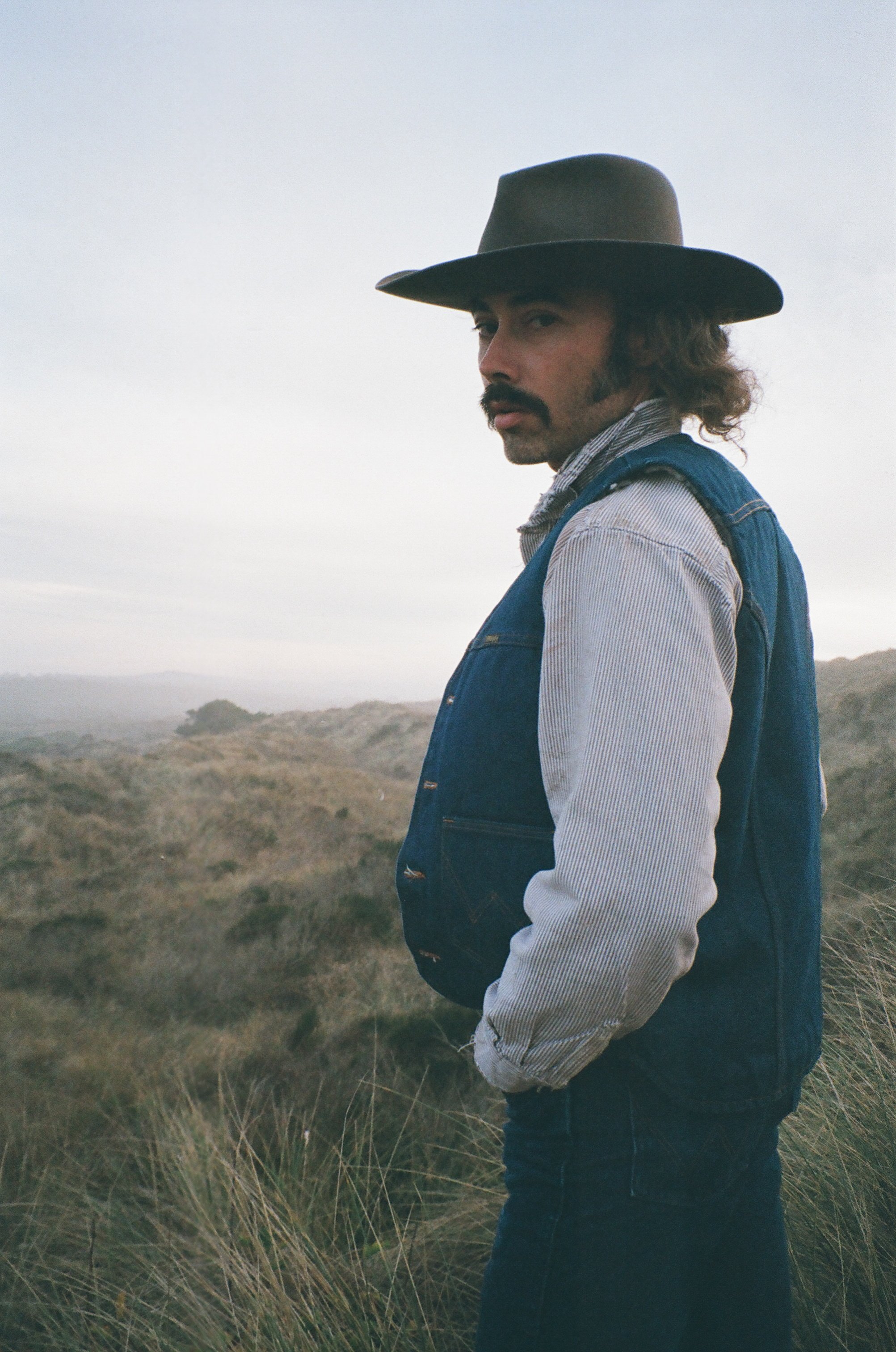 Man with long hair and mustache wearing a wide-brimmed hat, striped shirt, and denim vest standing outdoors in a grassy landscape.