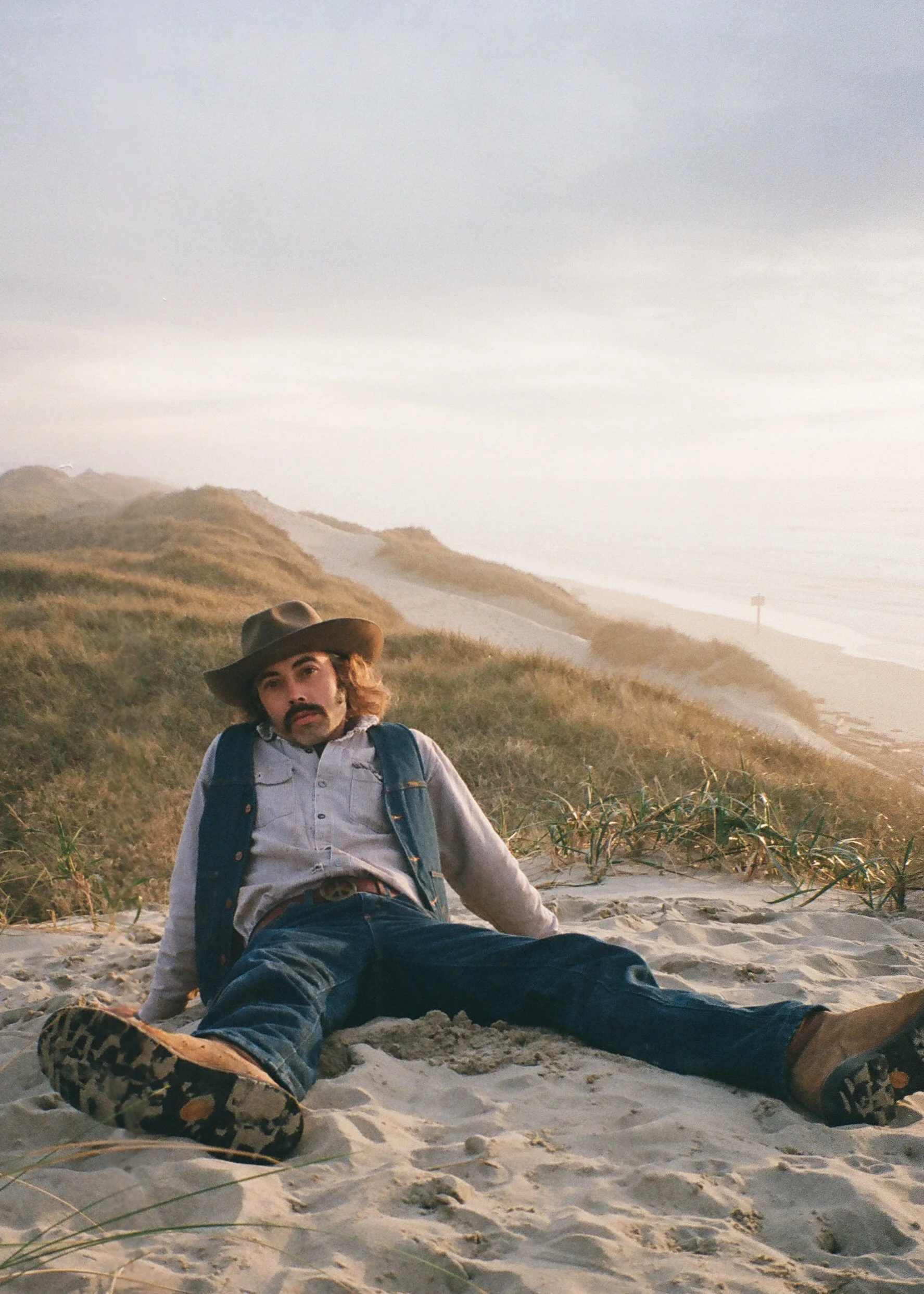 Man with long hair, mustache, and beard sitting on sandy beach dunes, wearing a cowboy hat, denim vest, light-colored shirt, jeans, and boots, with ocean and grassy dunes in the background.