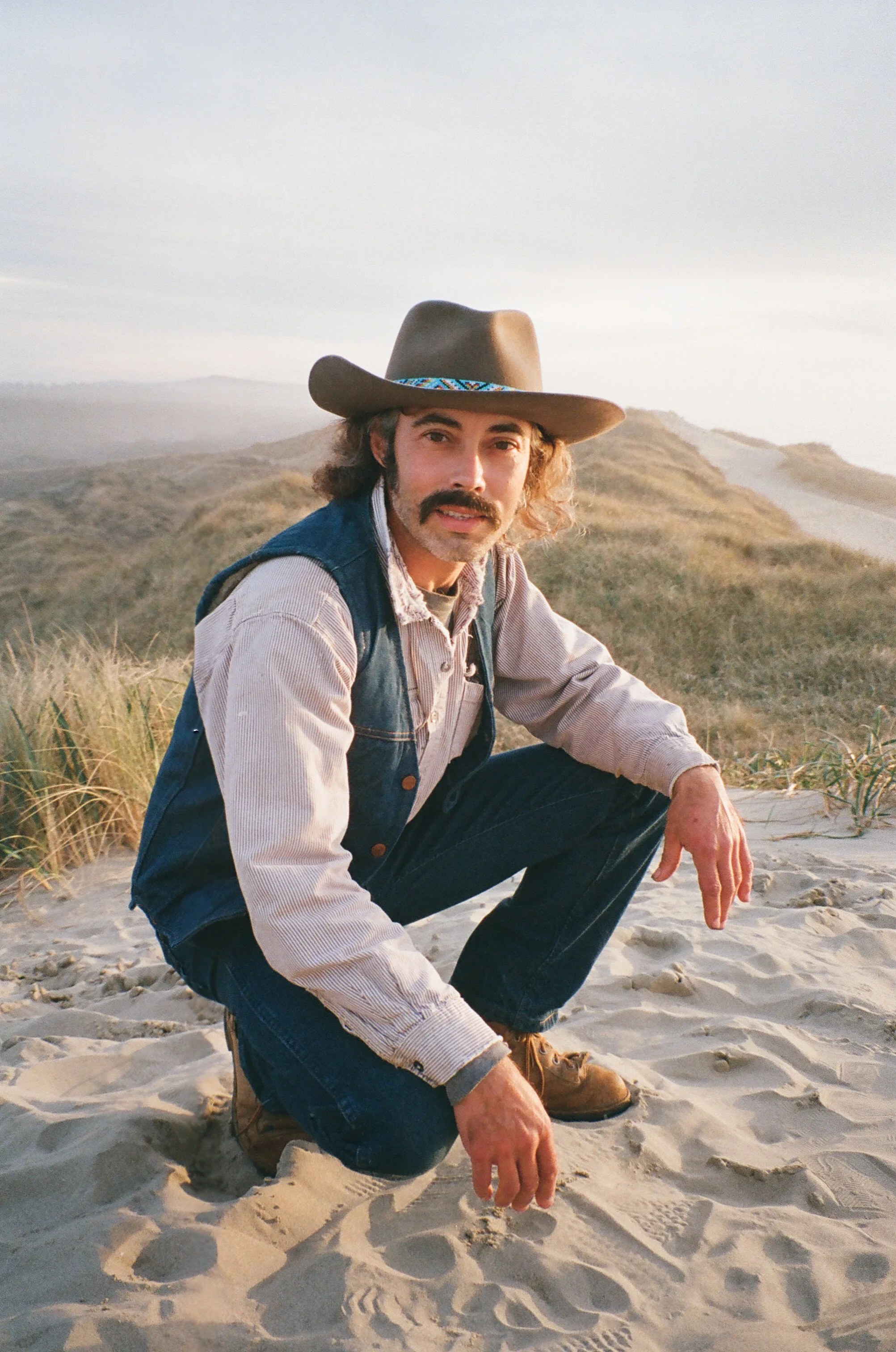 A man in a cowboy hat, denim vest, and jeans crouching on sandy terrain with grassy dunes and hills in the background during sunset.