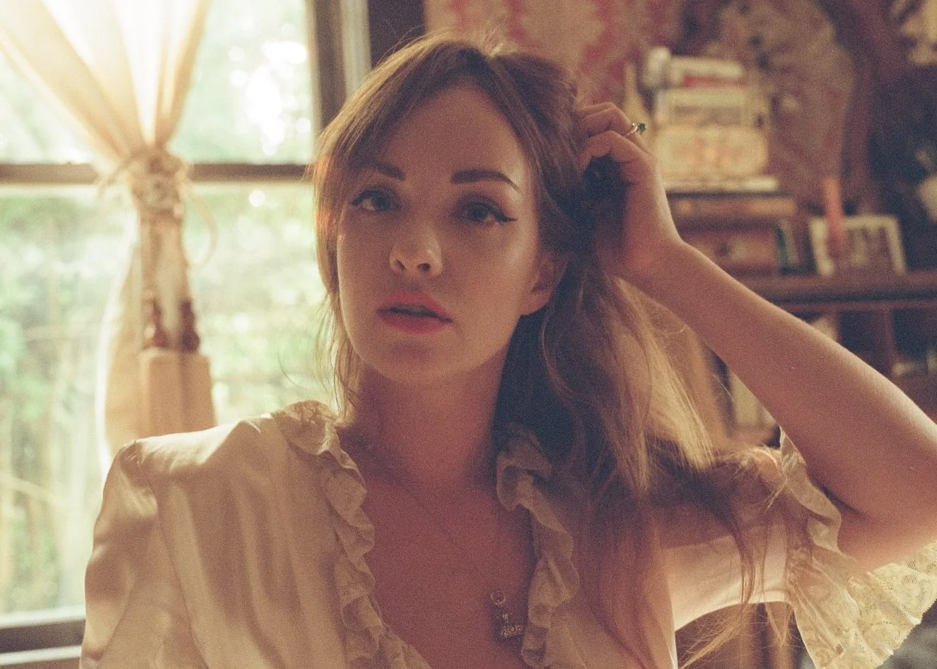 A young woman with wavy reddish-brown hair and fair skin, wearing a light-colored ruffled blouse and a necklace, sitting indoors with natural light coming through a window behind her, touching her hair with her left hand.