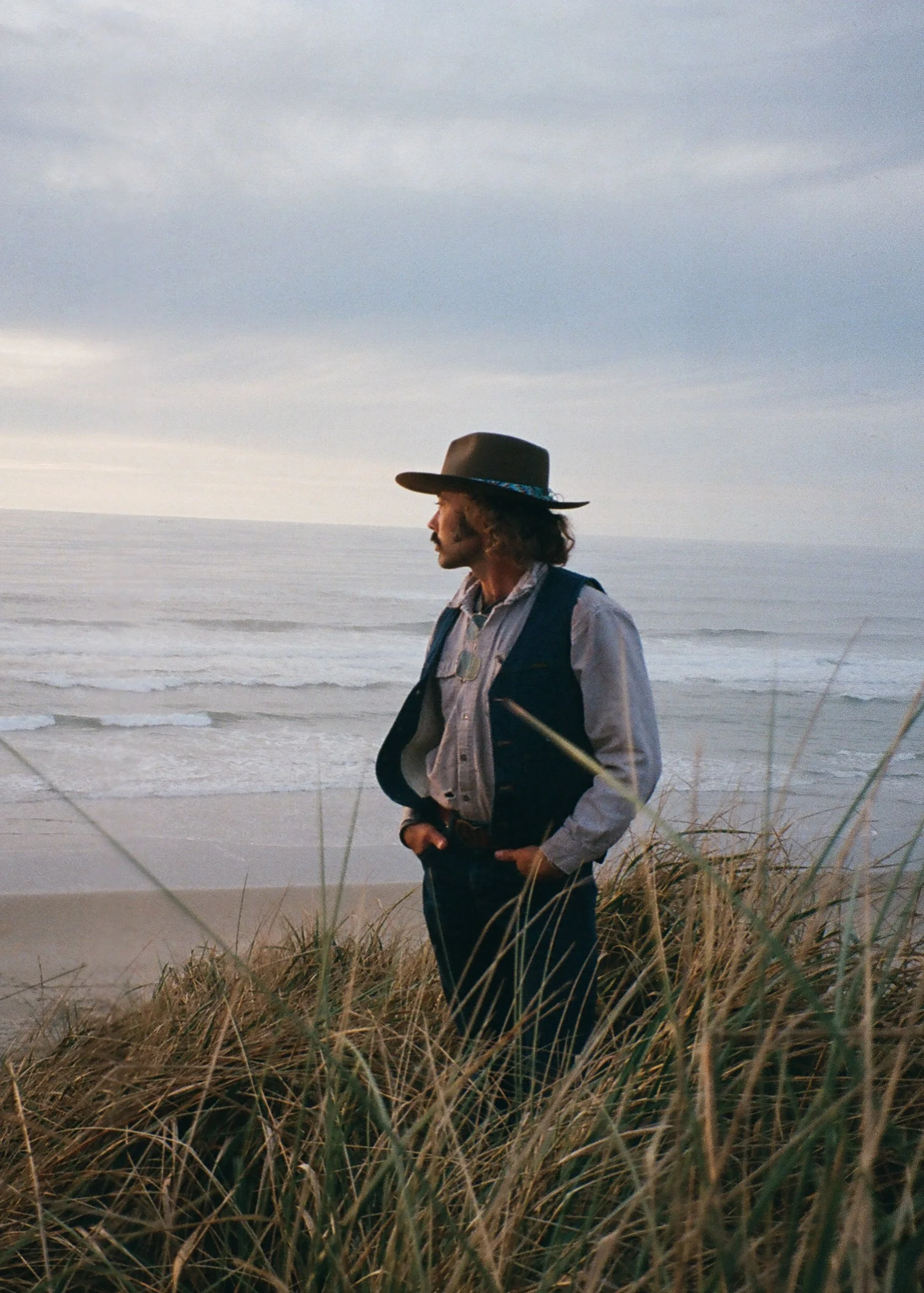 A man with shoulder-length hair and a beard, dressed in cowboy attire, stands on beach dunes with grass, gazing out at the ocean during sunset or sunrise.