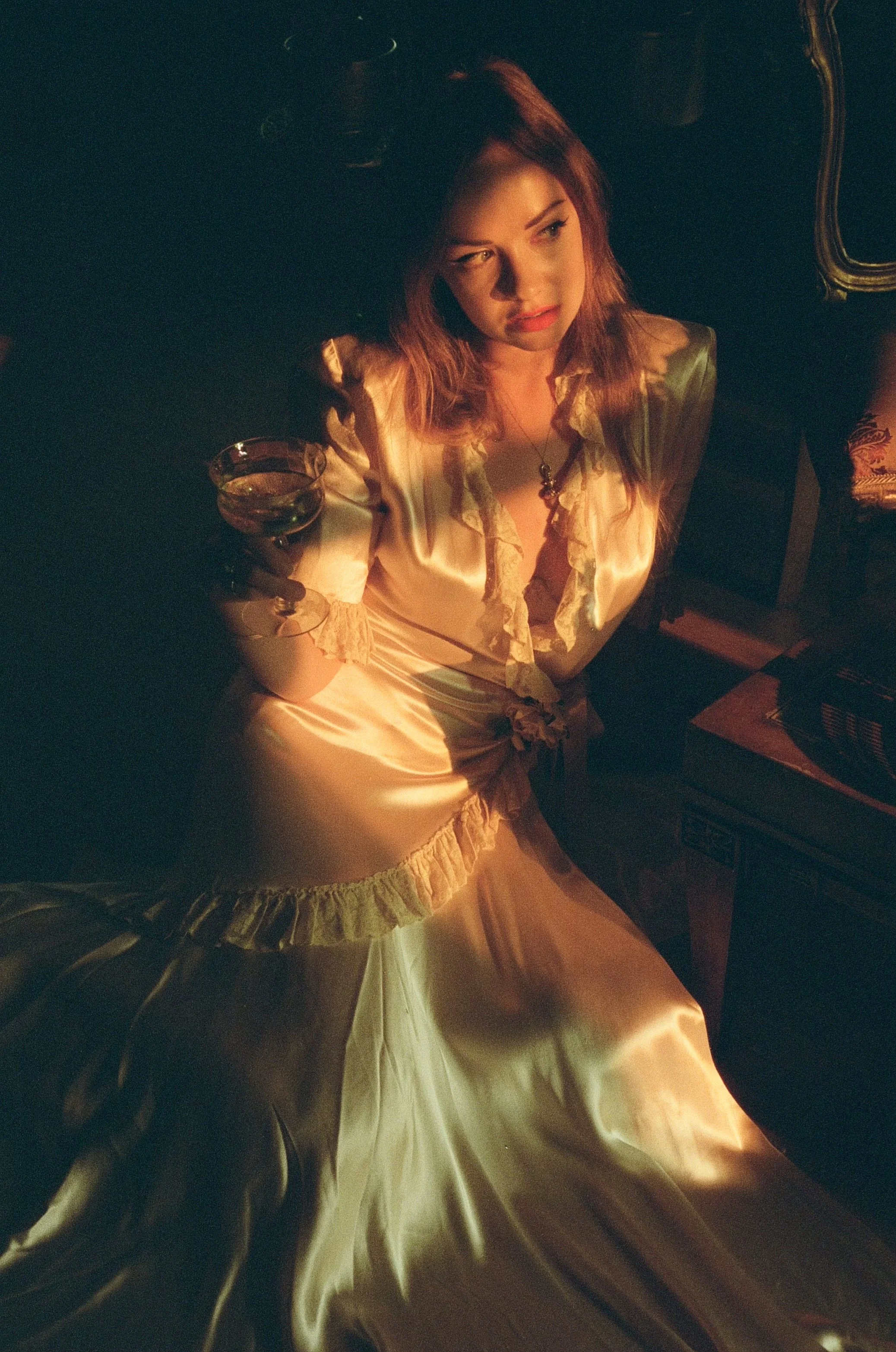 A woman with reddish hair sitting in a dimly lit room, wearing a shiny satin nightgown with ruffled edges, holding a glass of wine, looking contemplative.