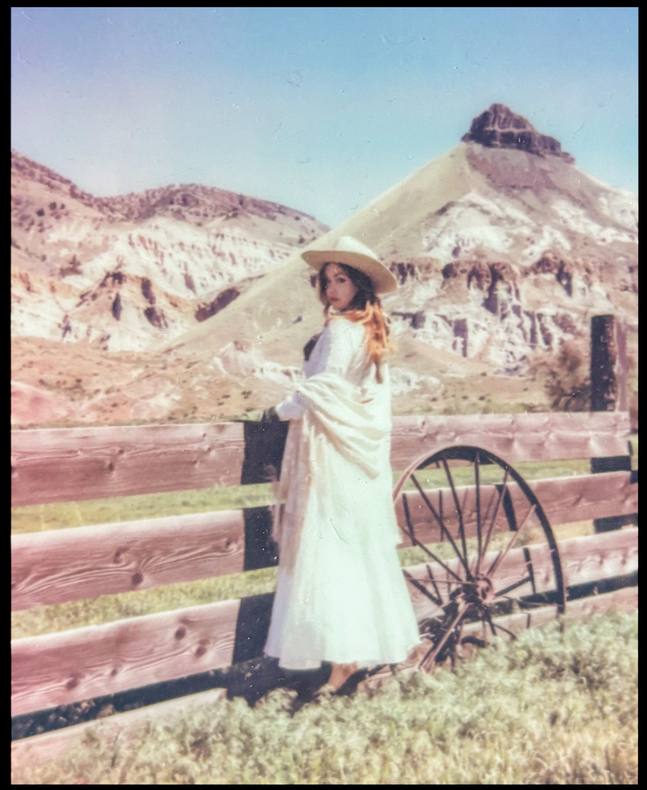A woman in a long white dress and a wide-brimmed hat standing by a wooden fence with a mountain and a hill in the background.