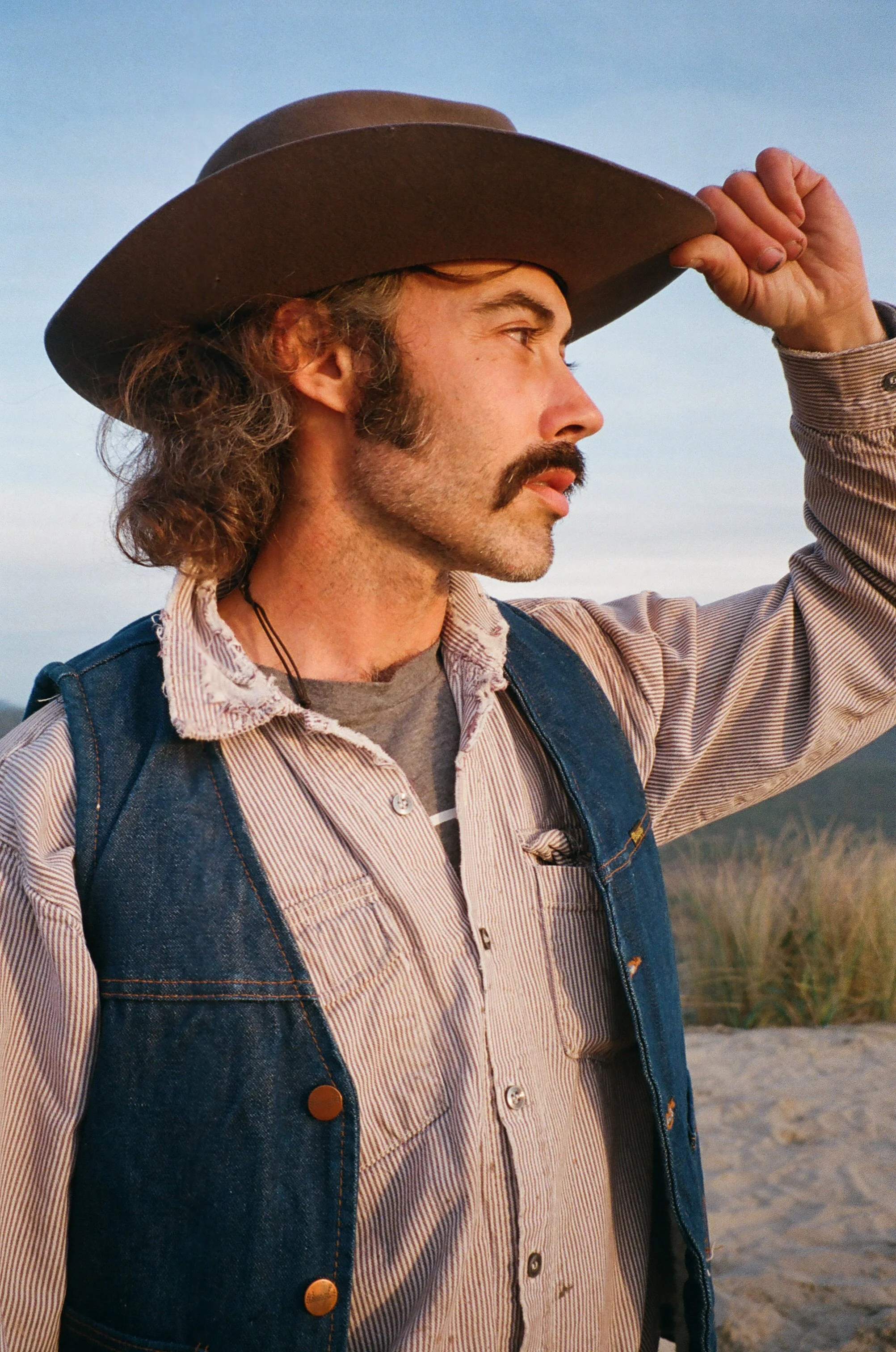 Profile of a man with long hair and mustache, wearing a cowboy hat, denim vest, and gray shirt, outdoors during sunset.
